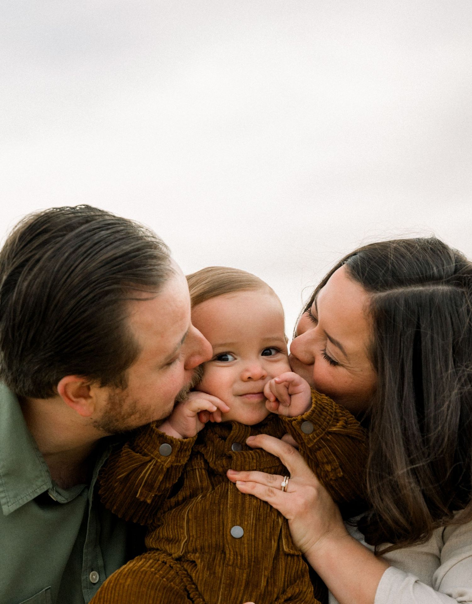 Parents lovingly squeeze and kiss their small child in brown jacket.