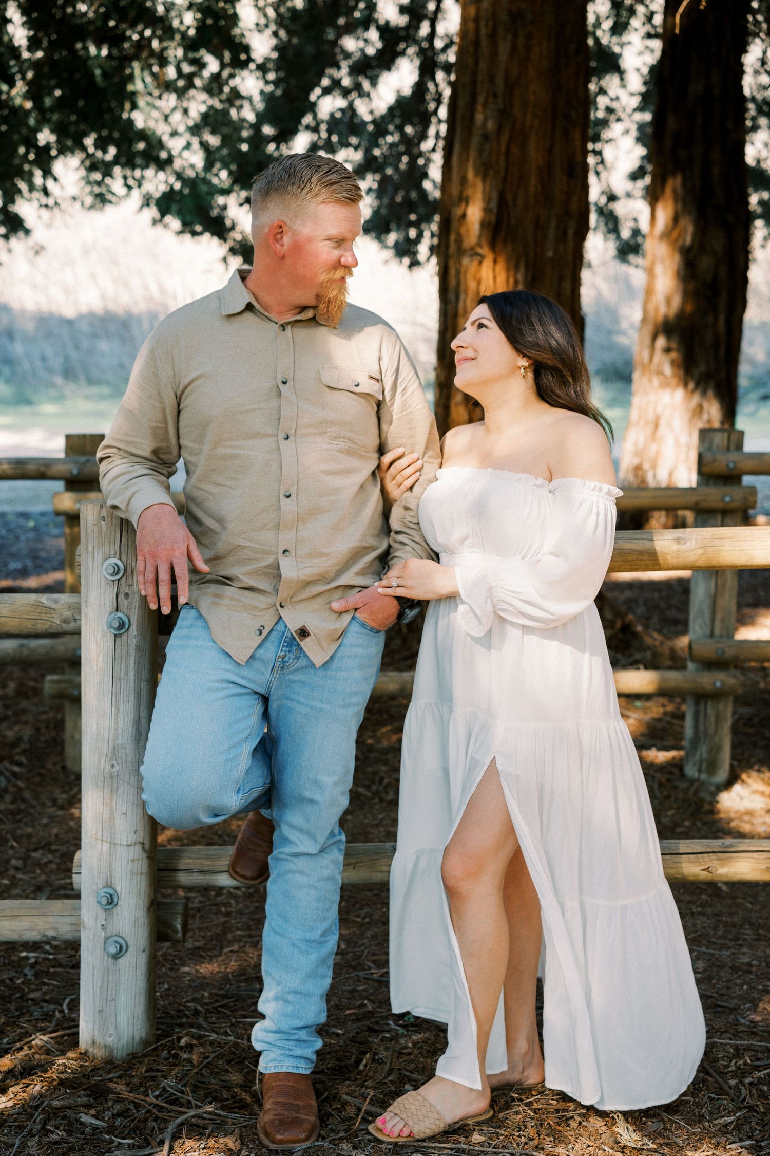 Couple poses together by wooden fence in outdoor setting with woman wearing flowing white dress.