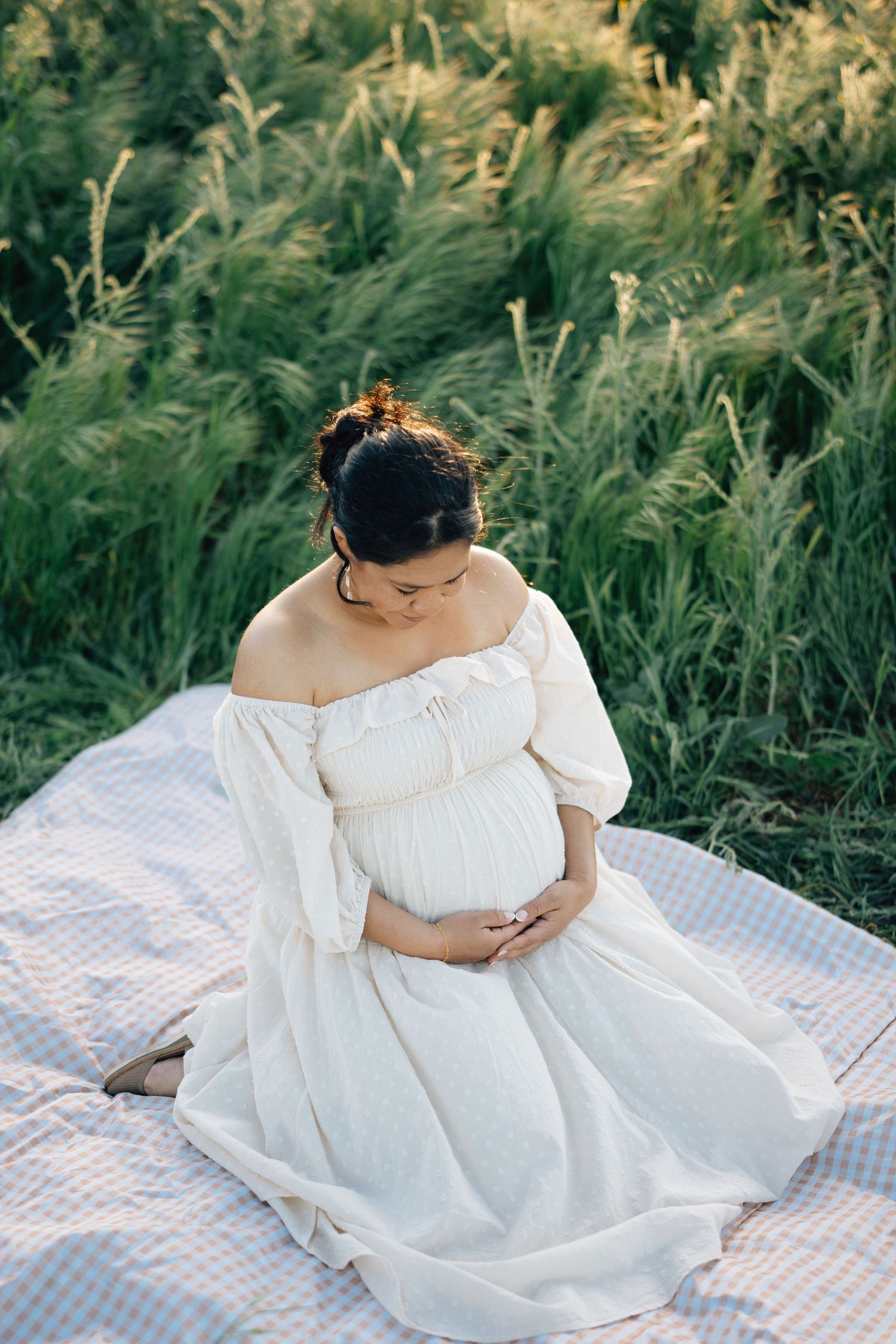 A pregnant woman in a white off-shoulder dress sits on a picnic blanket surrounded by tall grass at sunset.