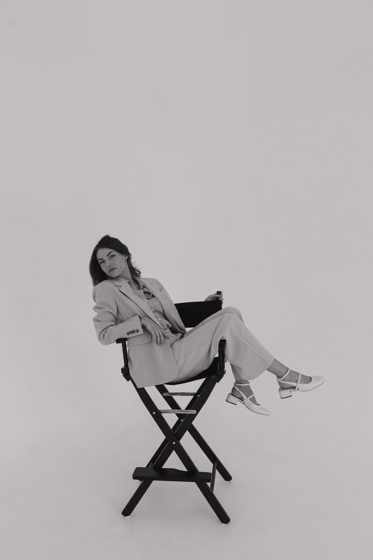Person relaxing in a black director's chair wearing a light colored outfit against a plain backdrop.