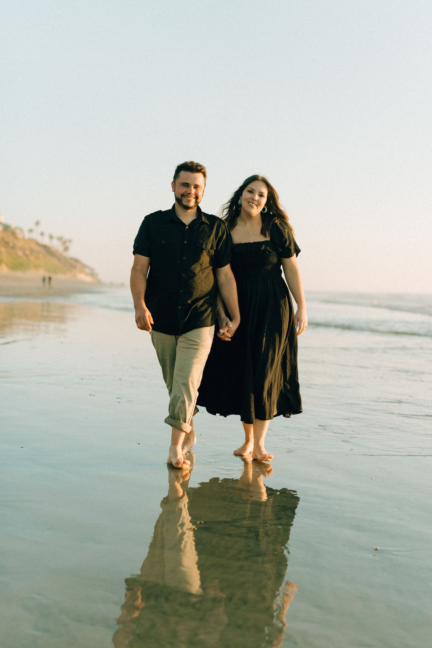 A couple walks hand in hand along the beach at sunset with waves reflecting their silhouettes.