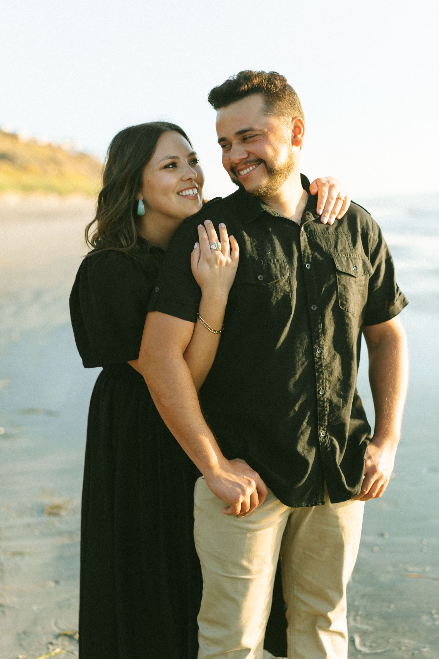 A couple shares a joyful moment on the beach while showing off an engagement ring.