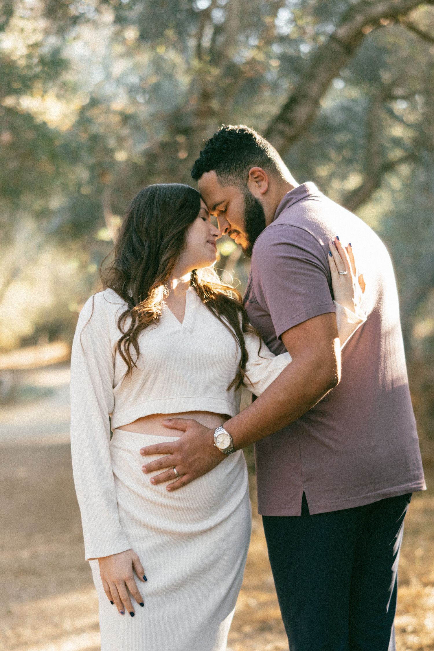 An expectant couple shares a romantic moment in a sunlit wooded area.