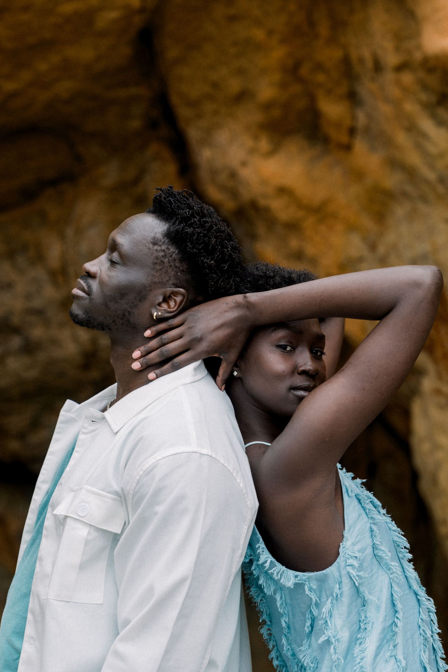 A stylish couple poses back-to-back against a rocky cave wall, wearing white and turquoise outfits.