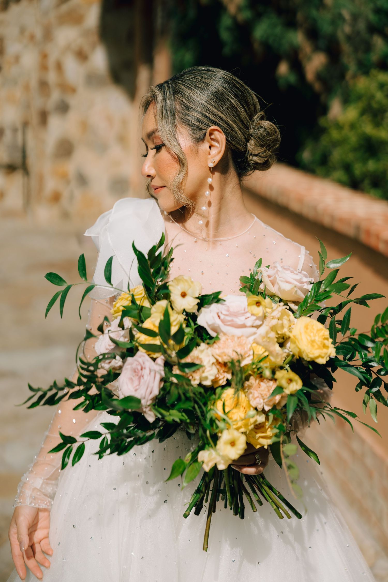 An elegant bridal portrait with a bouquet of yellow and pink flowers against a stone wall.