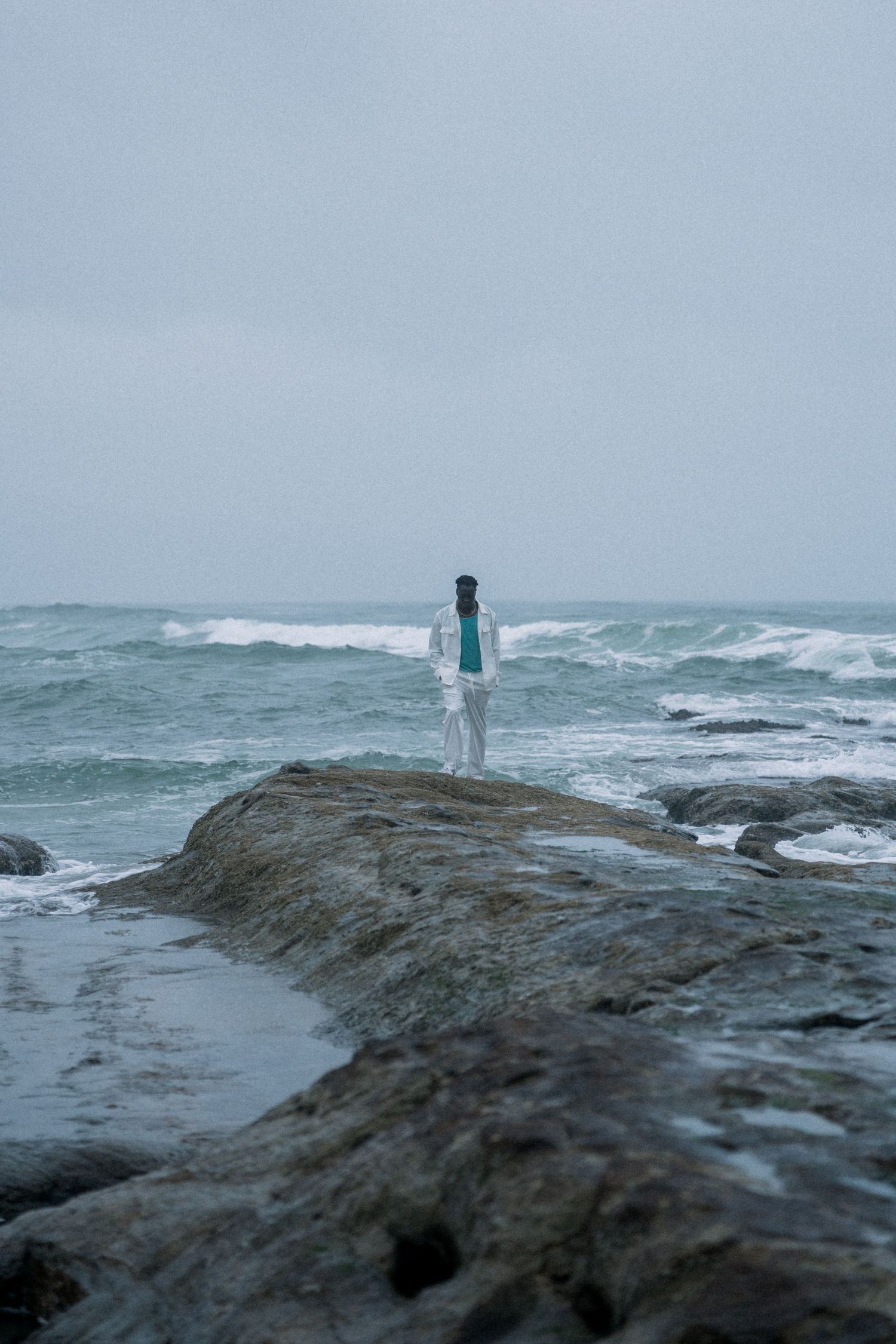 A solitary figure in white stands on rocky coastal cliffs overlooking stormy ocean waves.