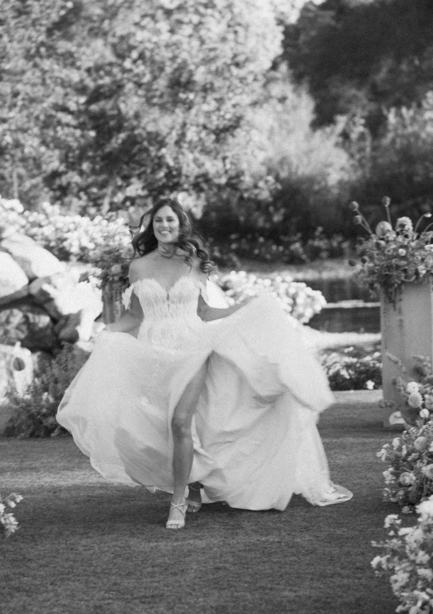 Candid black and white photo of bride dancing in flowing gown among garden flowers.