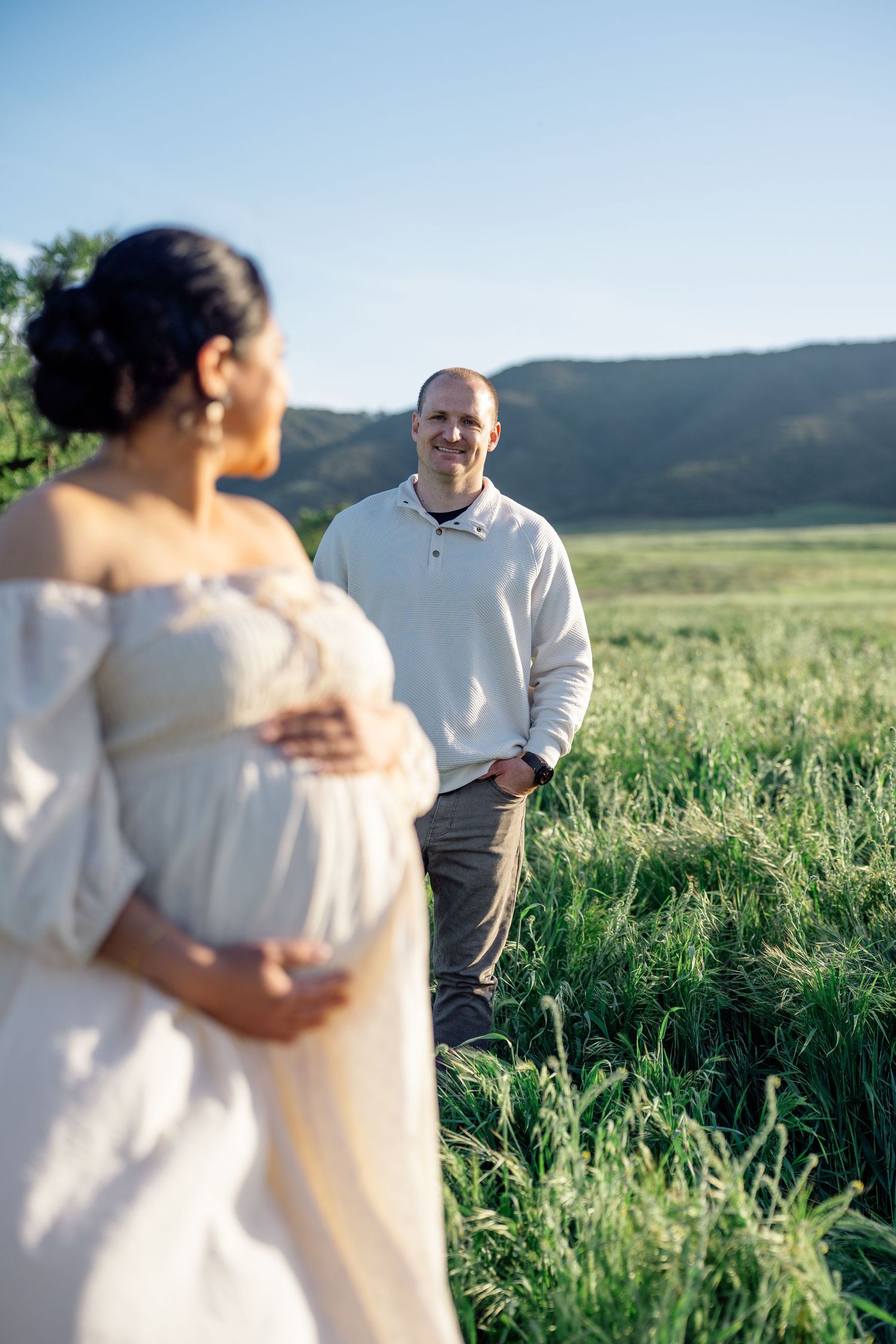 A couple standing in a green meadow with rolling hills, woman in white off-shoulder dress caressing her baby bump.