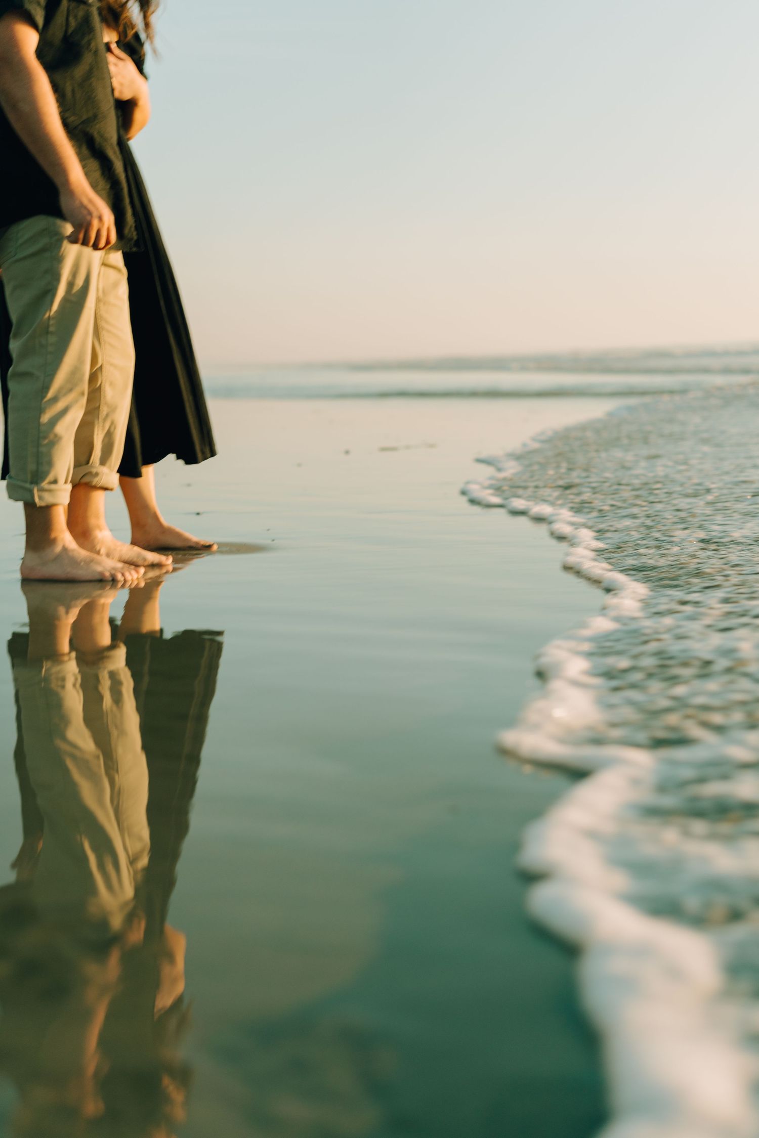 Cropped view of couple's feet reflected in wet sand at ocean's edge during sunset.
