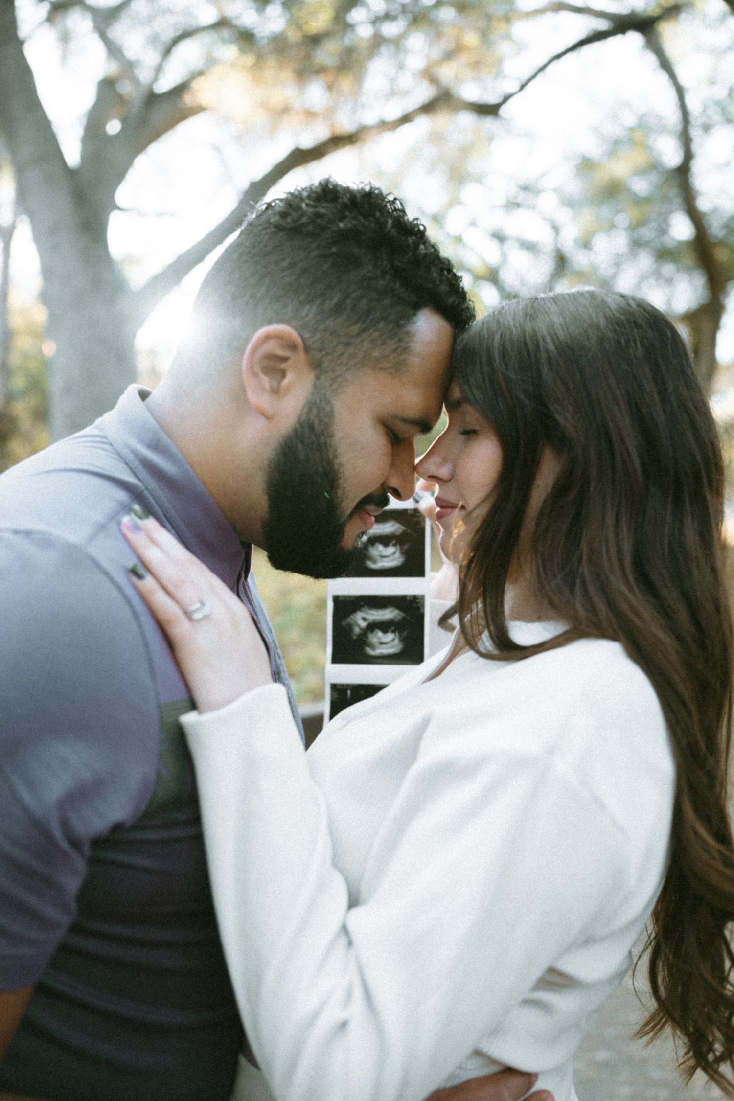 A couple shares an intimate moment while holding an ultrasound photo outdoors.