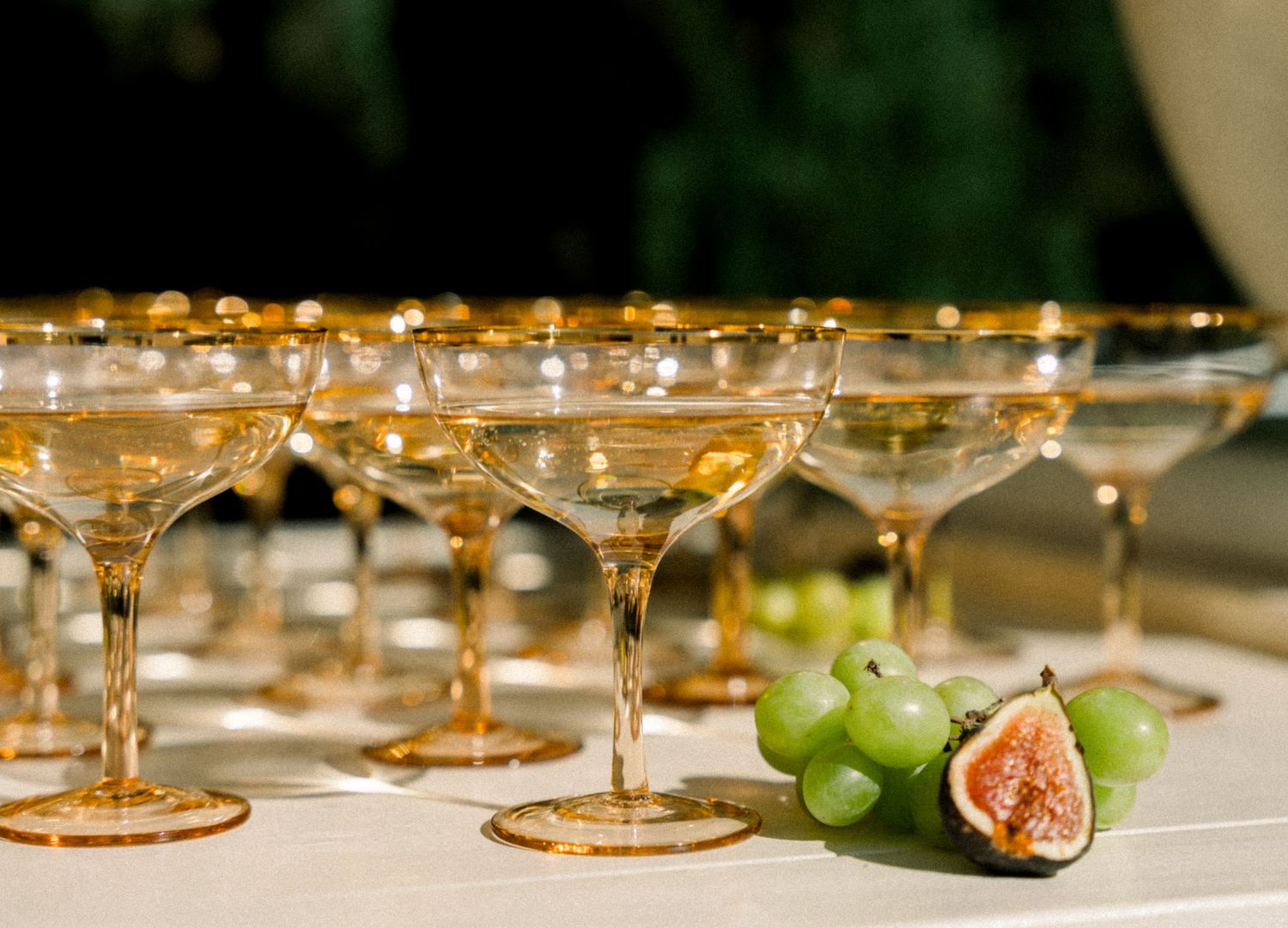Vintage gold-rimmed champagne coupes lined up with fresh figs and grapes as garnish.
