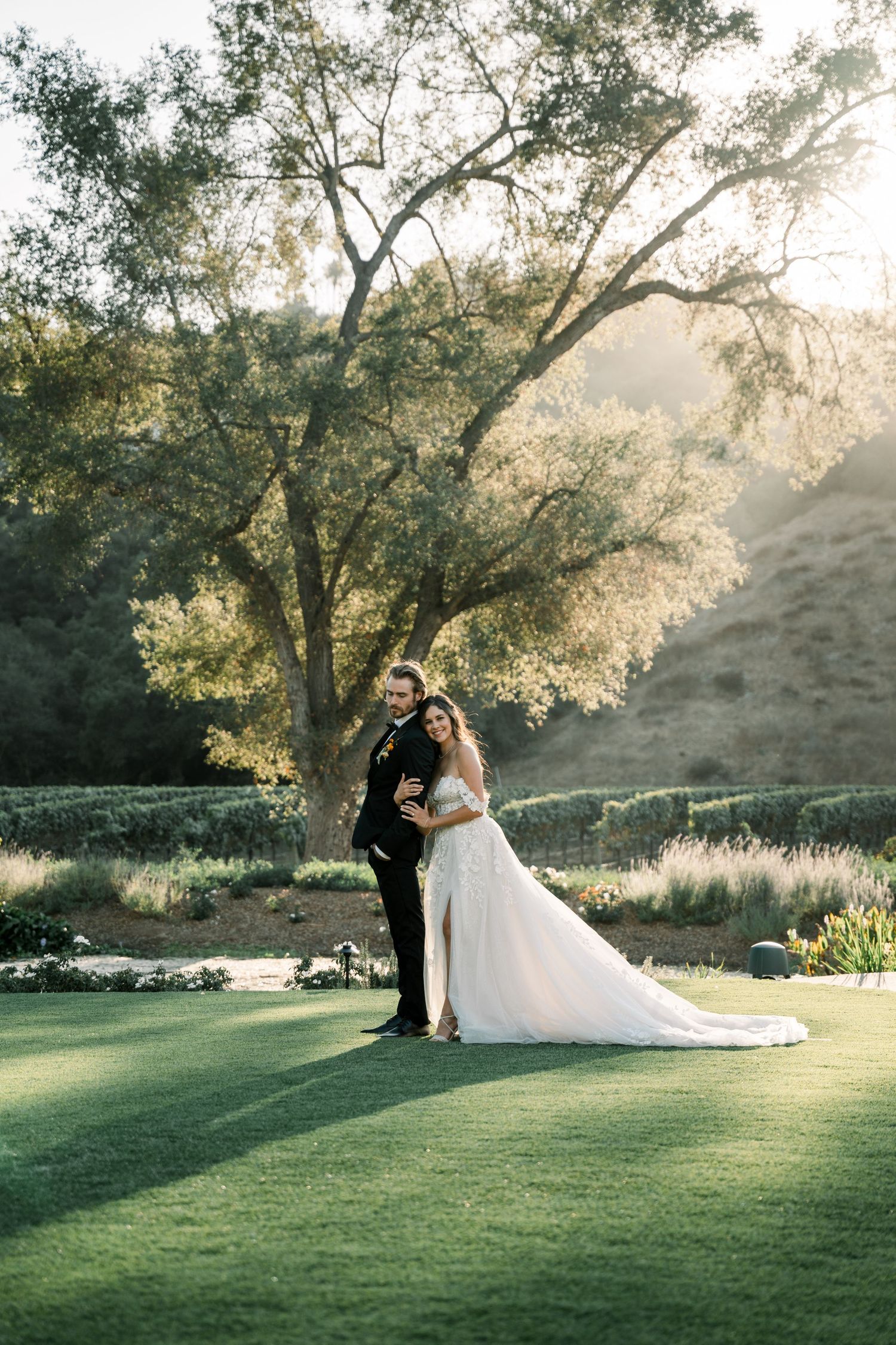 A couple in wedding attire embrace under a large tree on a manicured lawn with hills in the background at sunset.