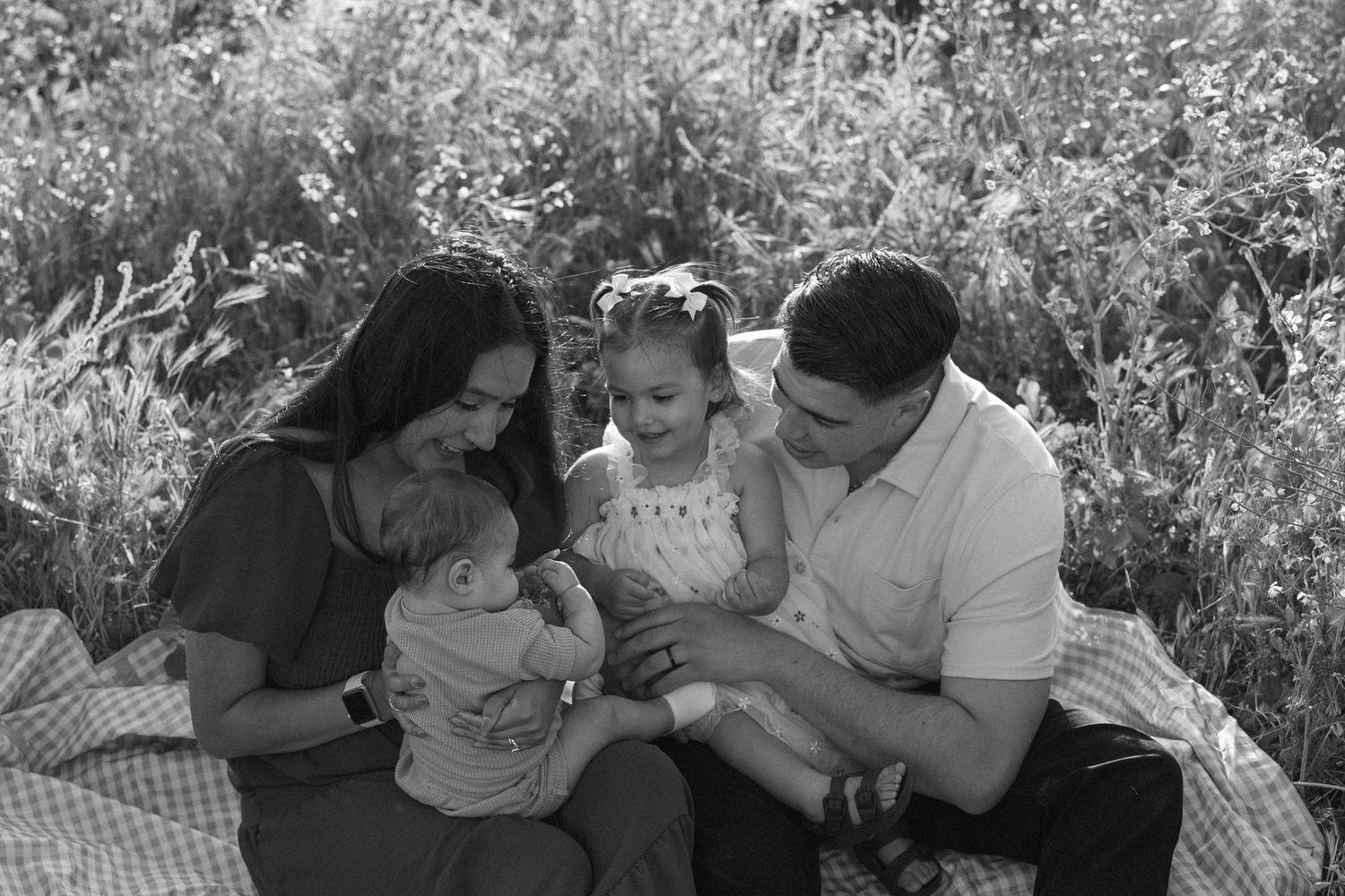 A family sits together on a blanket outdoors for a black and white portrait session surrounded by greenery.