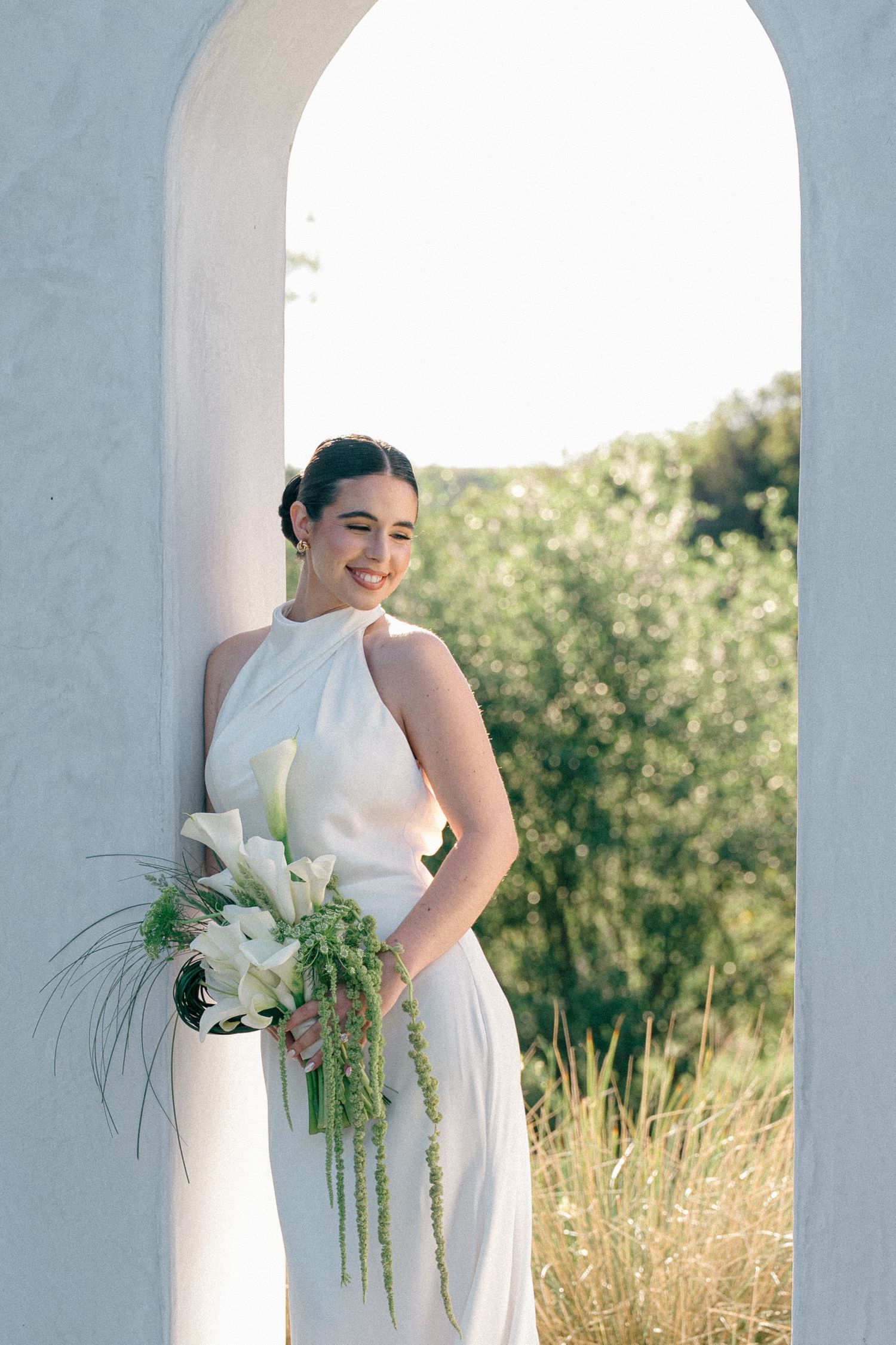 A bride in a white wedding dress poses elegantly against white arches with sunlit grass and greenery in the background.