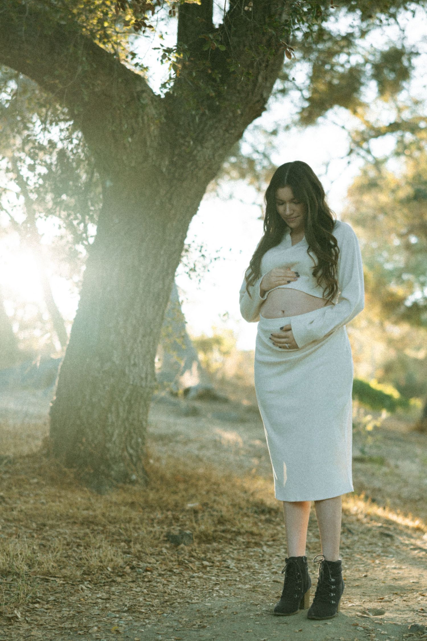 A woman in a white dress stands alone in dappled sunlight beneath a large tree.