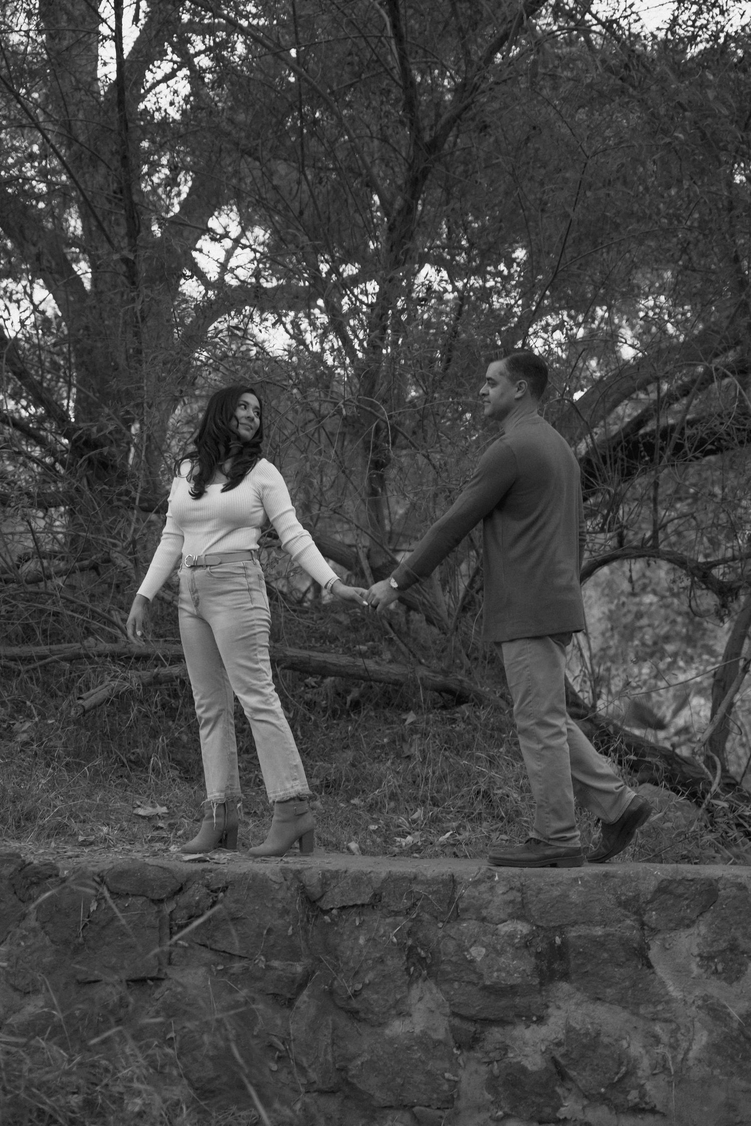 Couple stands together on stone wall in black and white engagement photo.