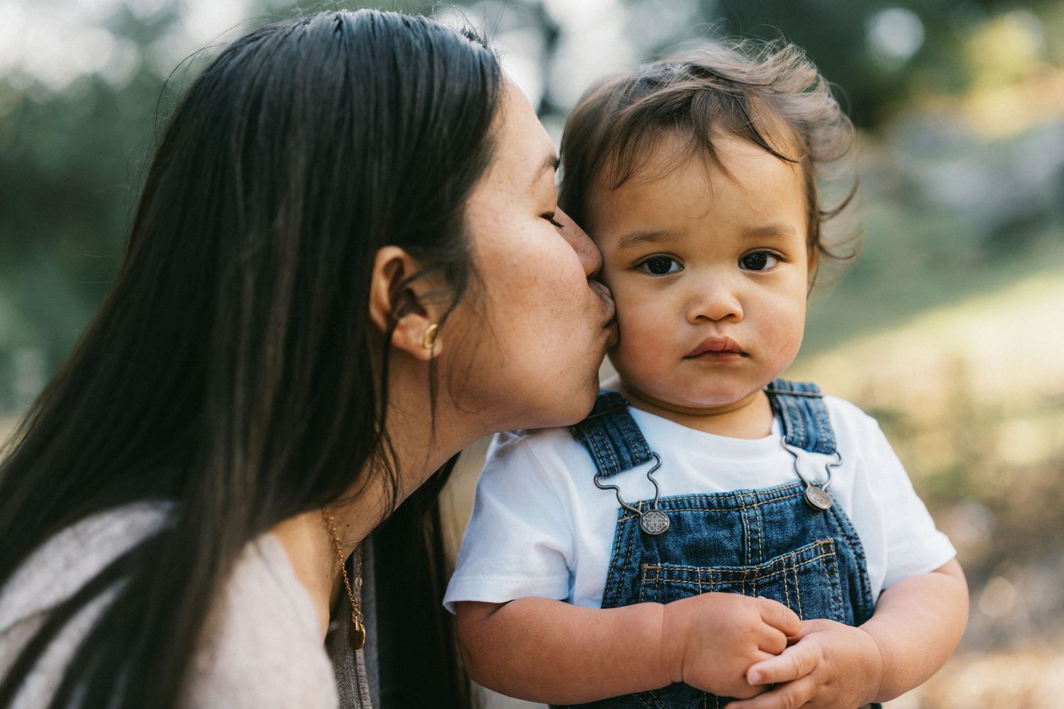 A parent lovingly kisses their baby wearing blue denim overalls while spending time together outdoors.