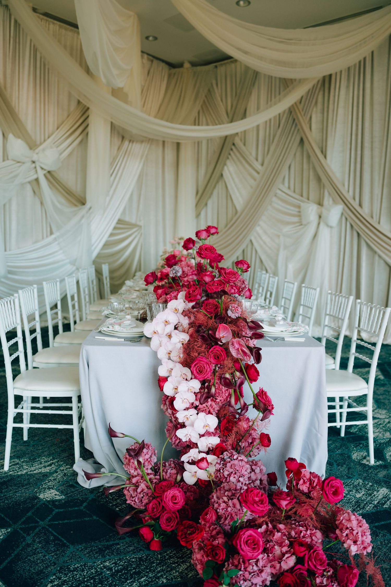 Elegant wedding reception table with dramatic red floral runner and white draped backdrop.