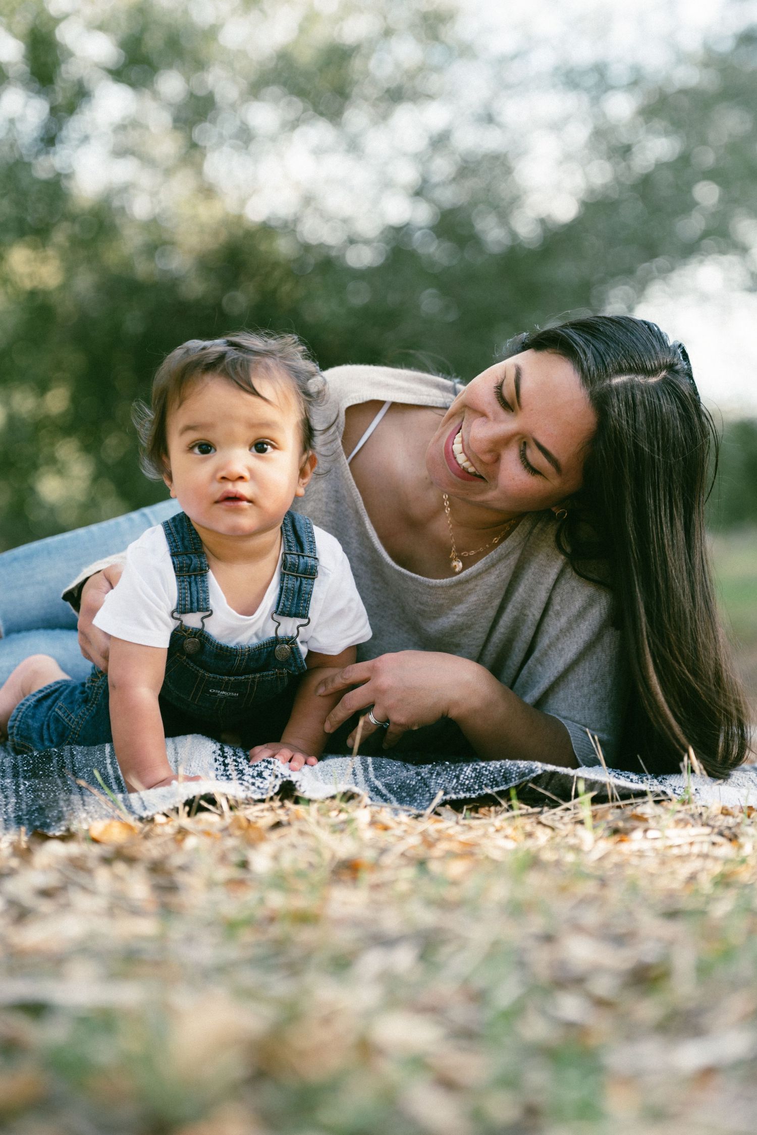Mother and baby in overalls share a joyful moment while lying on fallen leaves in an outdoor setting.