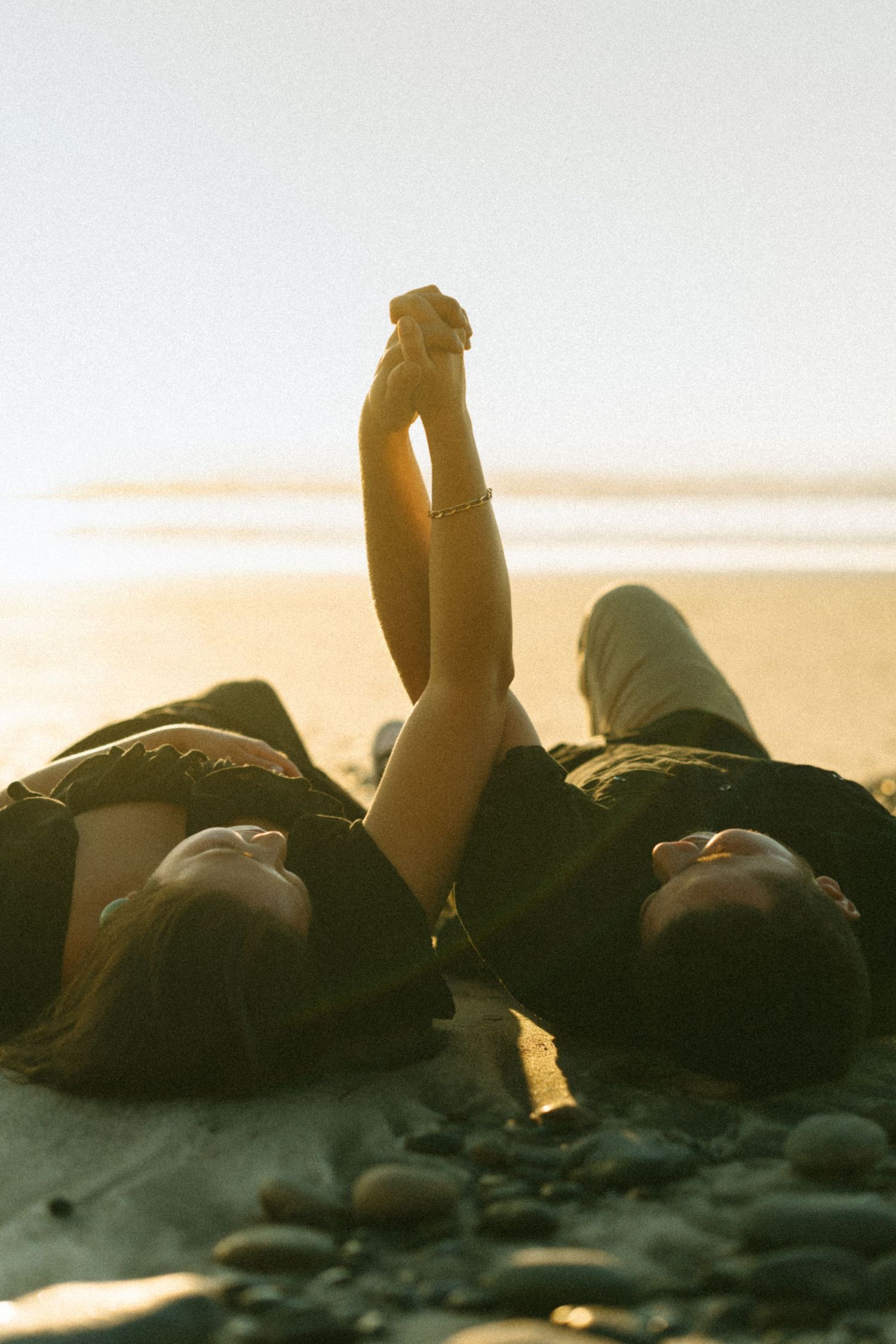 Two people lying on beach holding hands up against sunset sky.