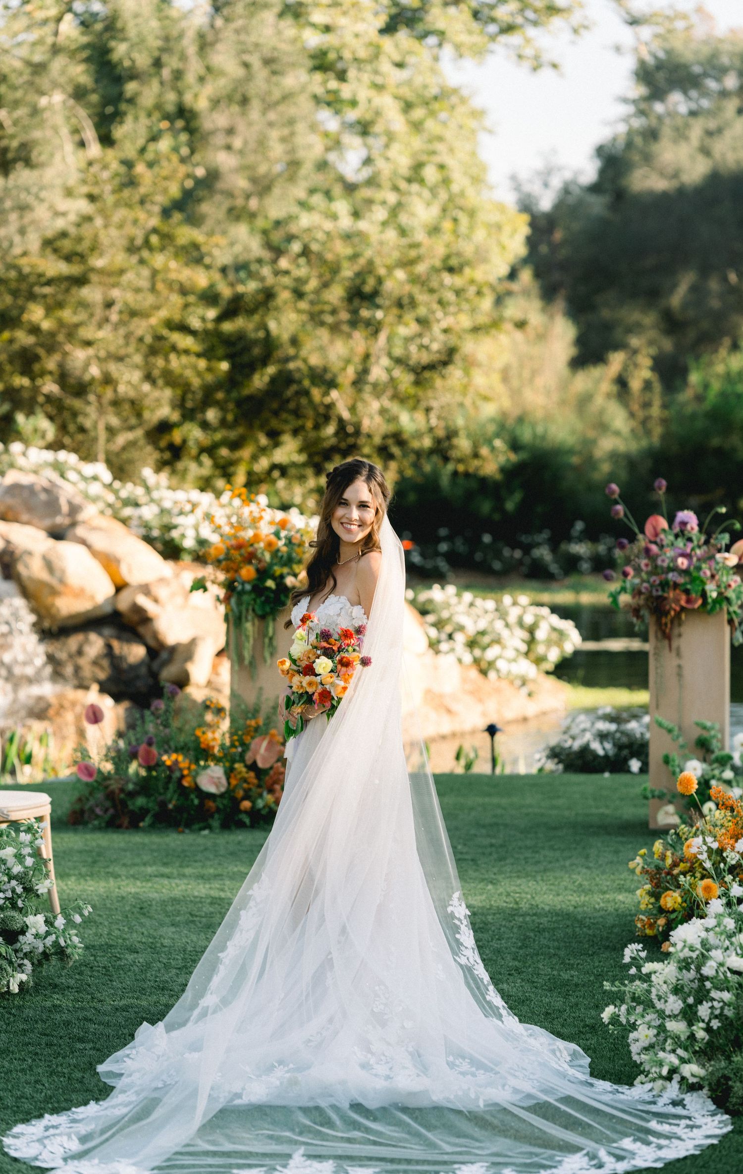 A wedding scene in a garden with flowing white veil and floral arrangements against lush greenery.