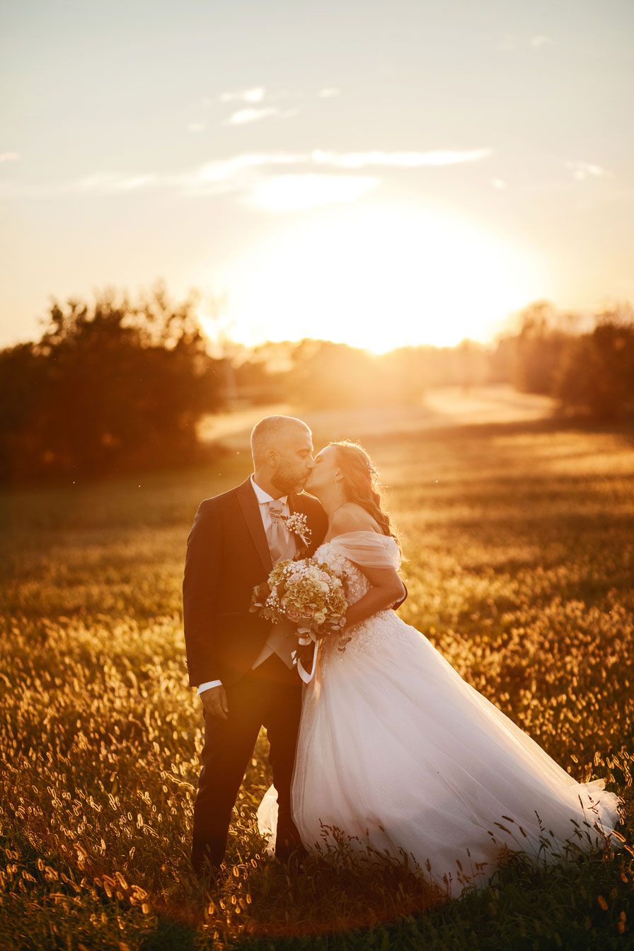 Romantica passeggiata al tramonto: gli sposi mano nella mano tra i campi, avvolti dalla magica luce della golden hour. Un'atmosfera calda e