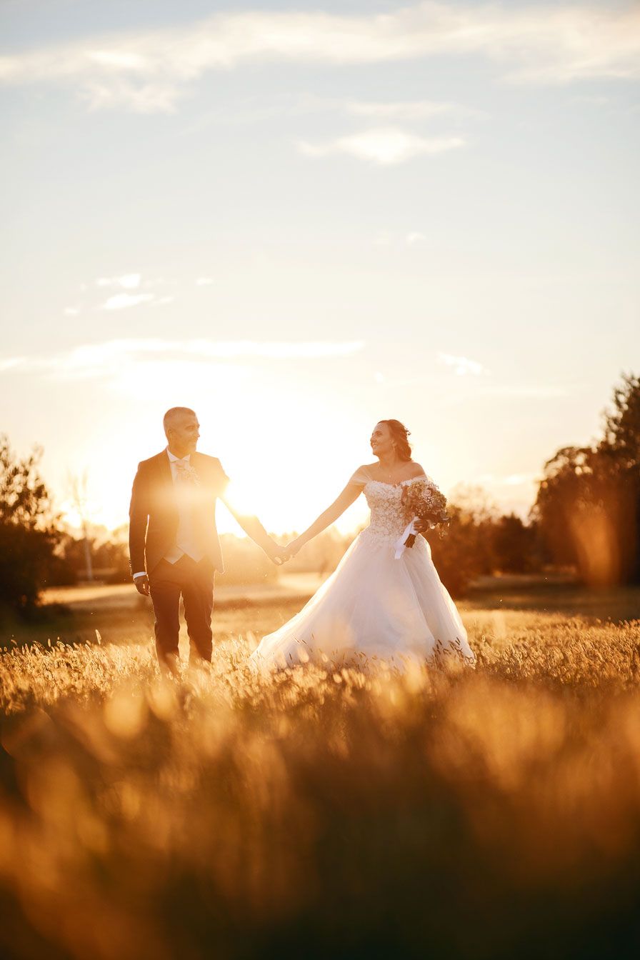 Romantica passeggiata al tramonto: gli sposi mano nella mano tra i campi, avvolti dalla magica luce della golden hour. Un'atmosfera calda e