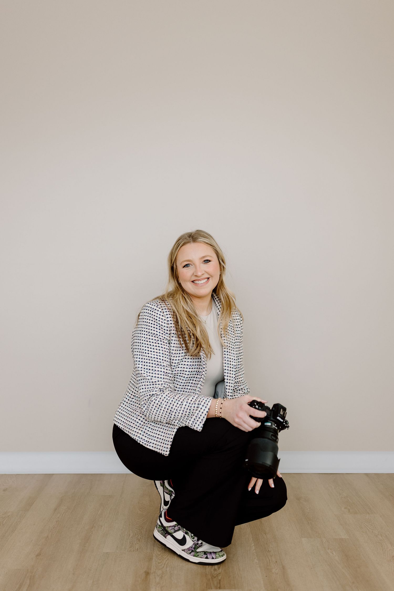 Smiling blonde woman crouching on wood floor holding a camera, wearing a houndstooth blazer and Nike sneakers.