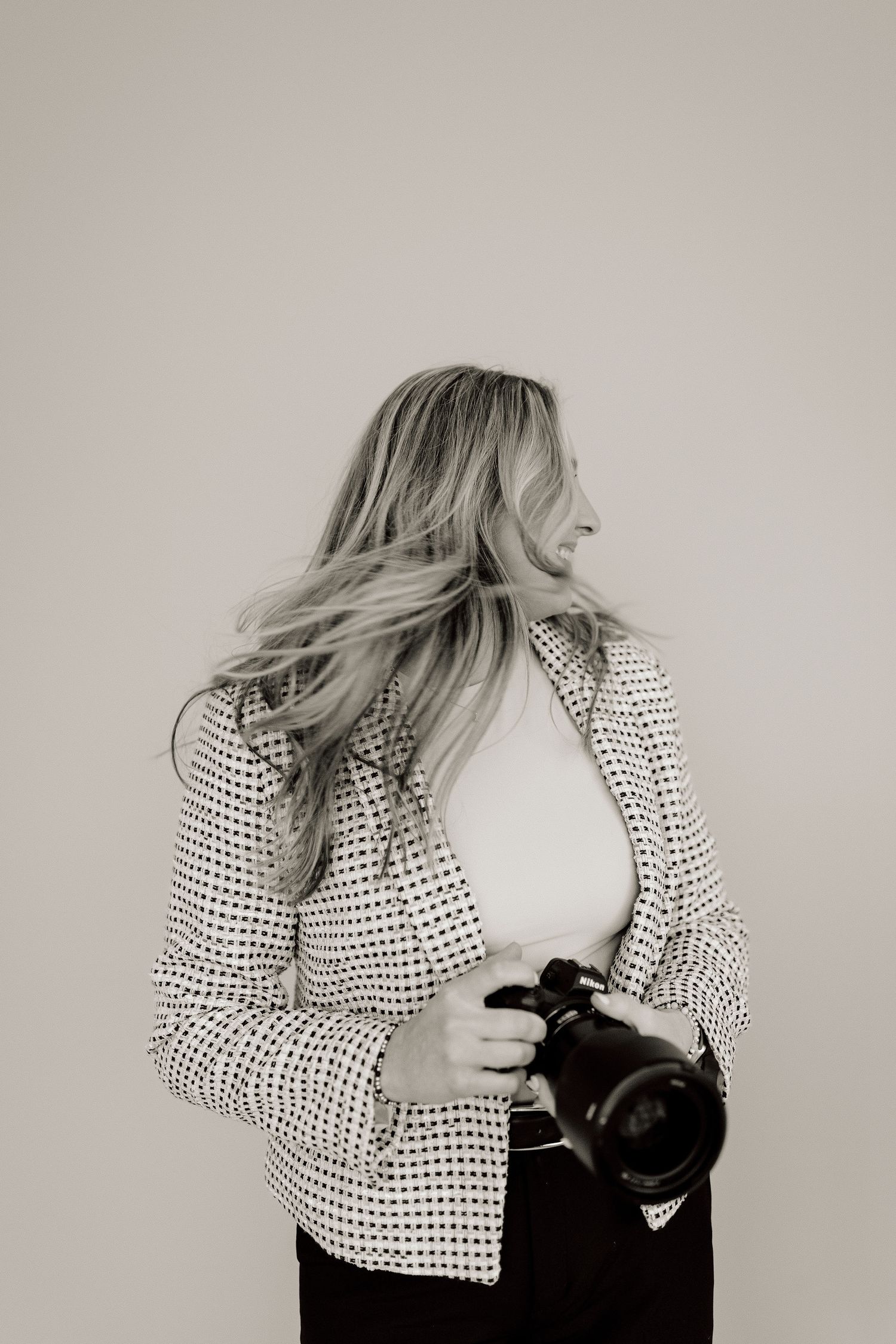 Black and white photo of a smiling blonde woman holding a camera, wearing a patterned blazer.