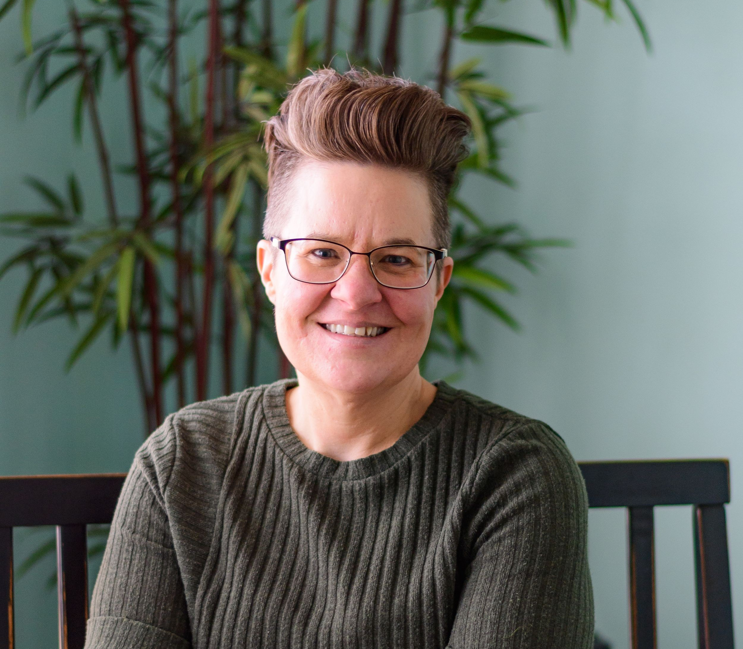 A person with glasses and a styled haircut smiles warmly while sitting in front of a bamboo plant backdrop.