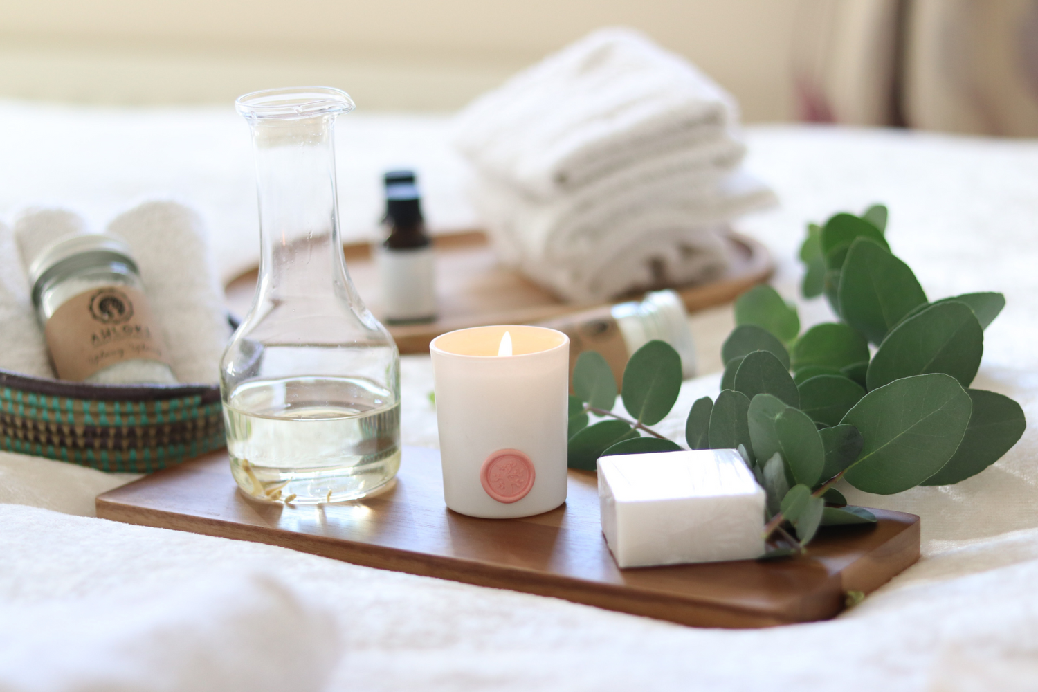 Spa amenities arranged on wooden tray with candle, towels, and eucalyptus leaves.