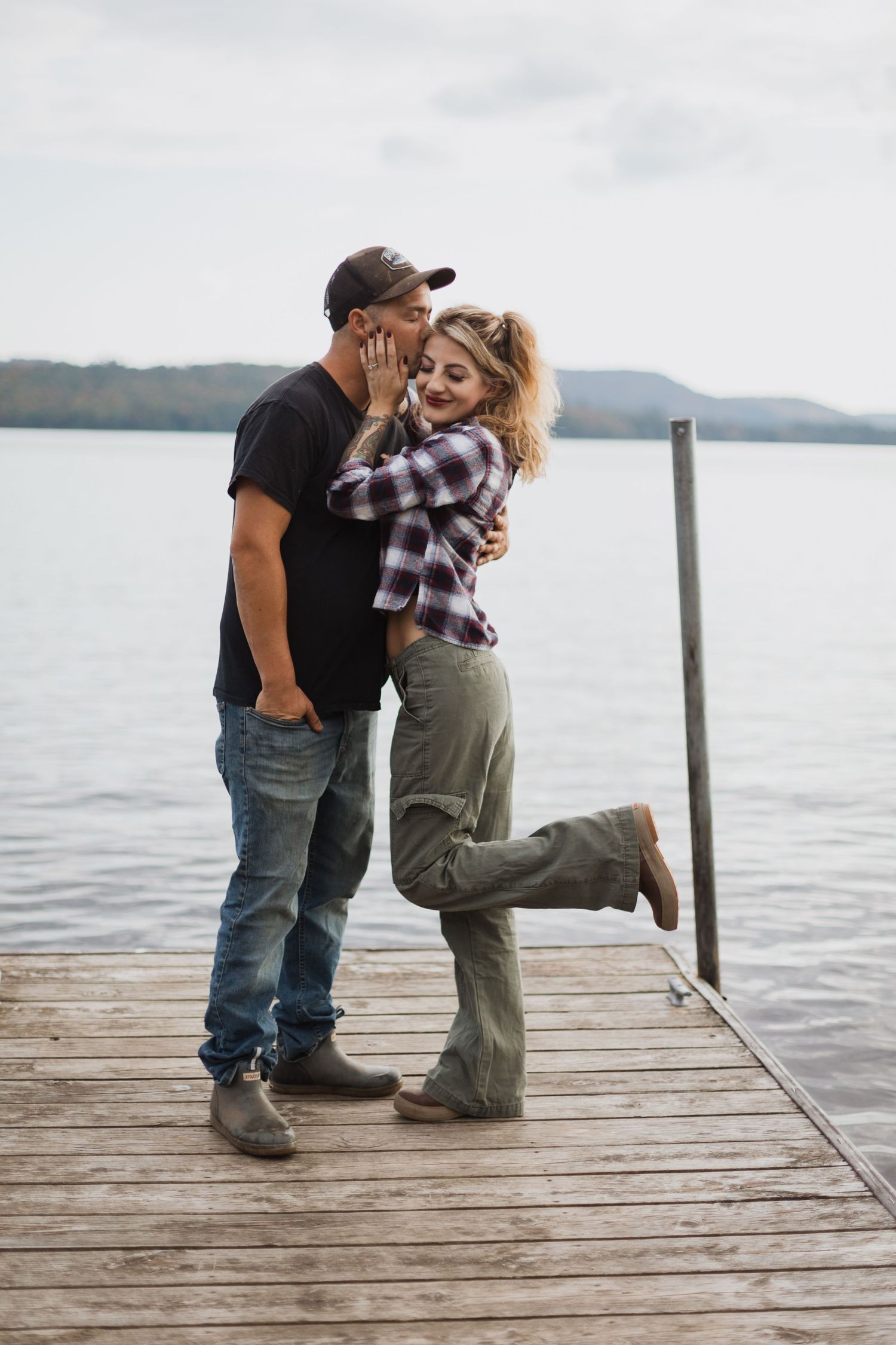 A playful couple shares a tender moment on a wooden pier with a lake and mountains behind them.