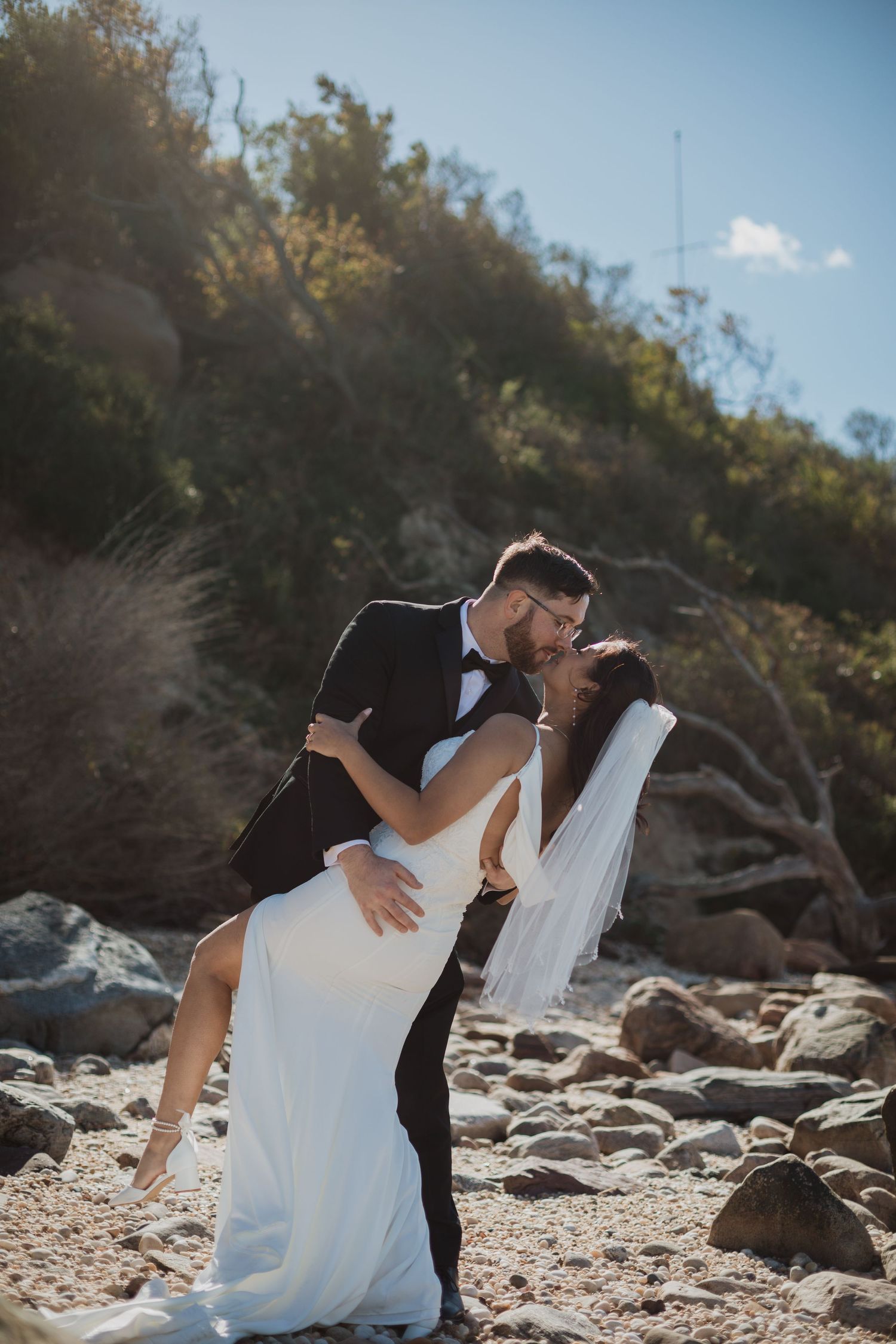 A dramatic dip kiss between newlyweds on a rocky beach with cliffs in the background.