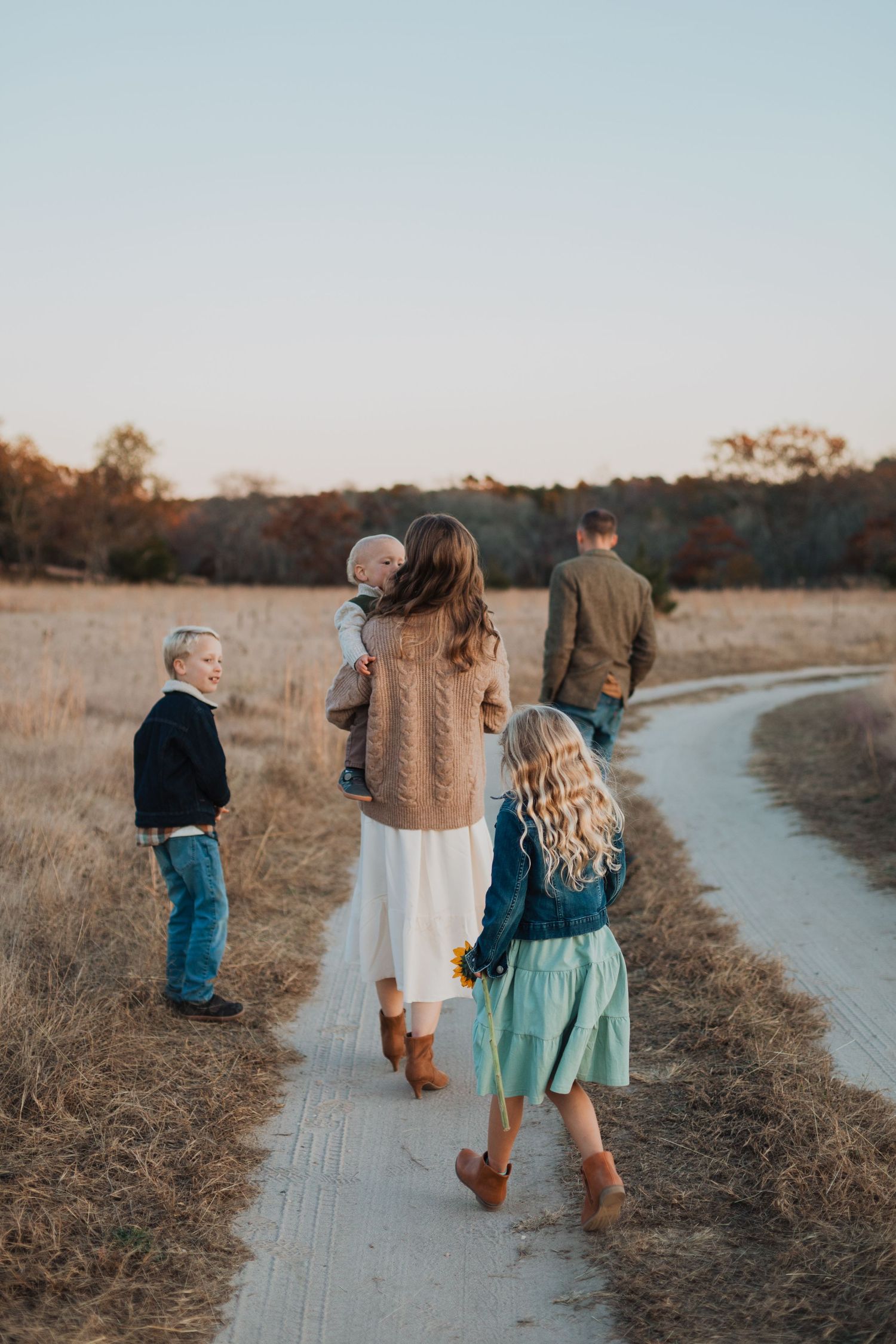 A family walks together on a winding dirt path through golden autumn fields at sunset.