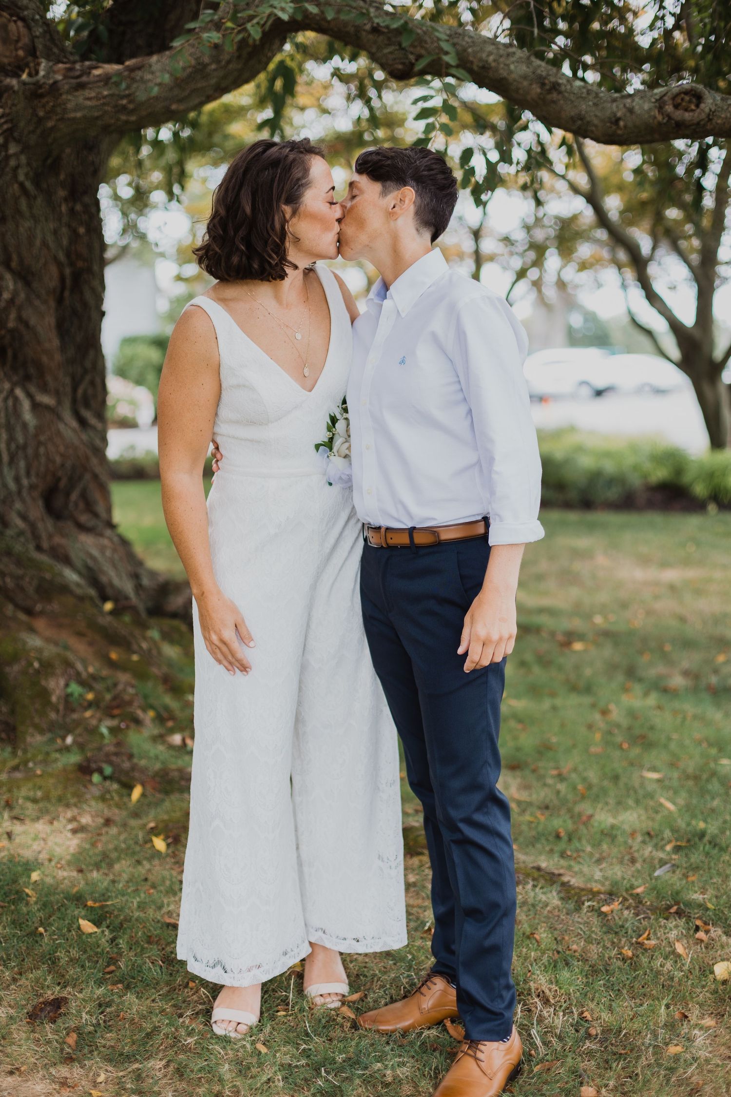 A couple in wedding attire shares intimate moments outdoors under trees on a grassy lawn.