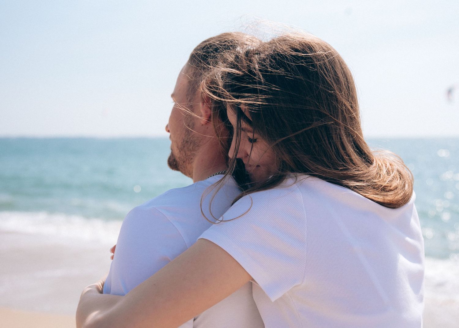A romantic couple shares an intimate moment together on a sunny beach with sparkling ocean waves in the background.