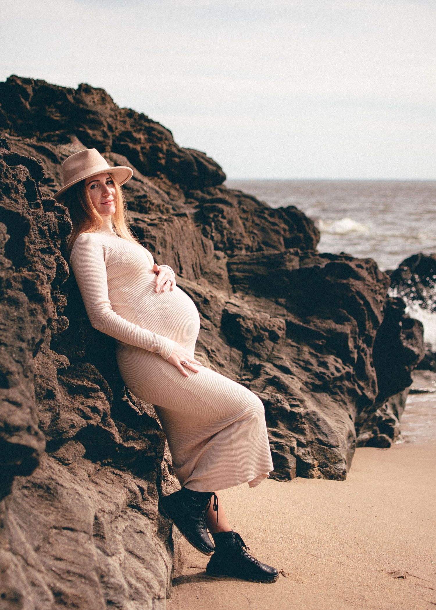 An expectant mother in a beige dress poses against rocky coastal cliffs during sunset.