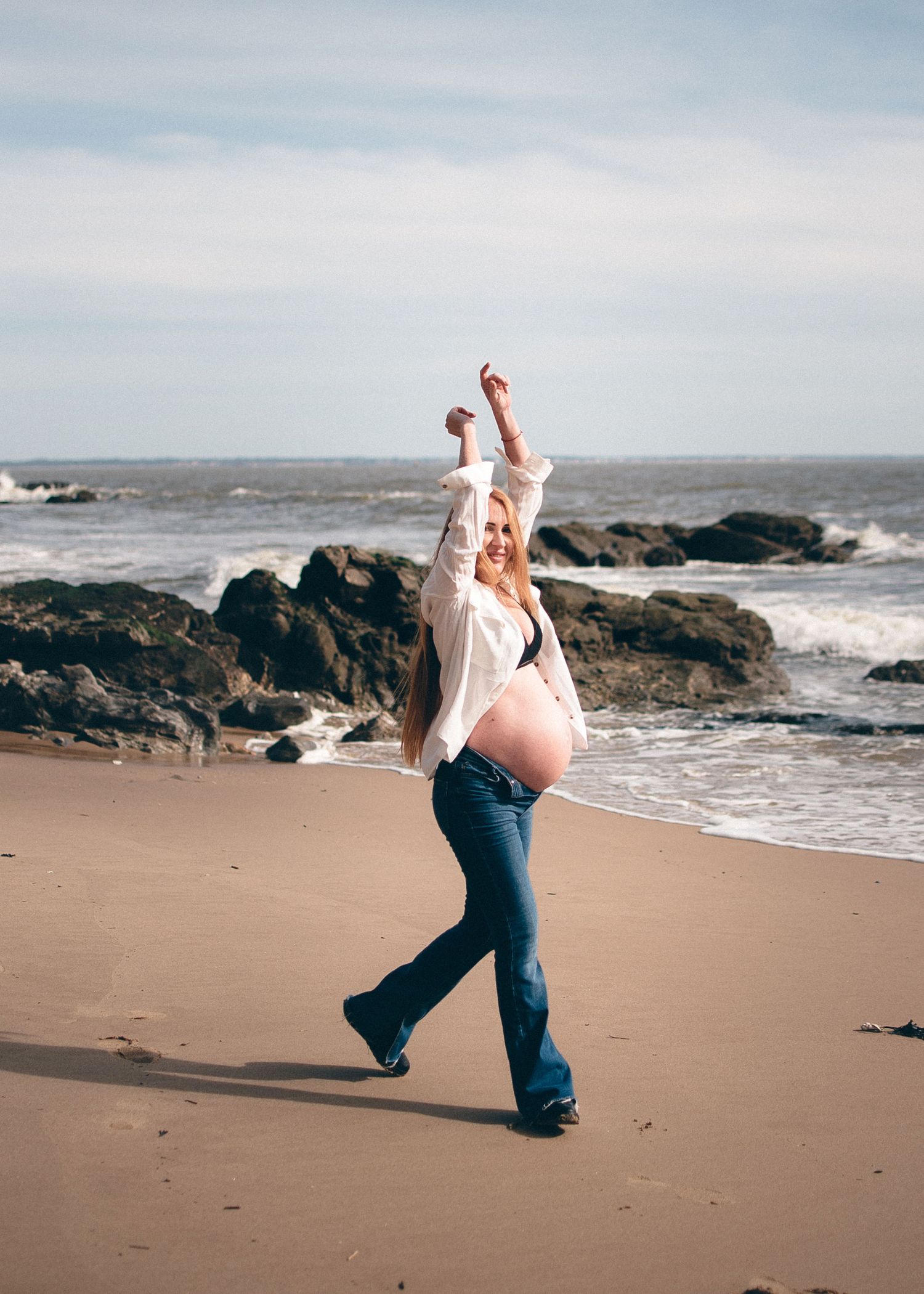 Person dancing joyfully on a beach with rocky coastline and waves in the background.