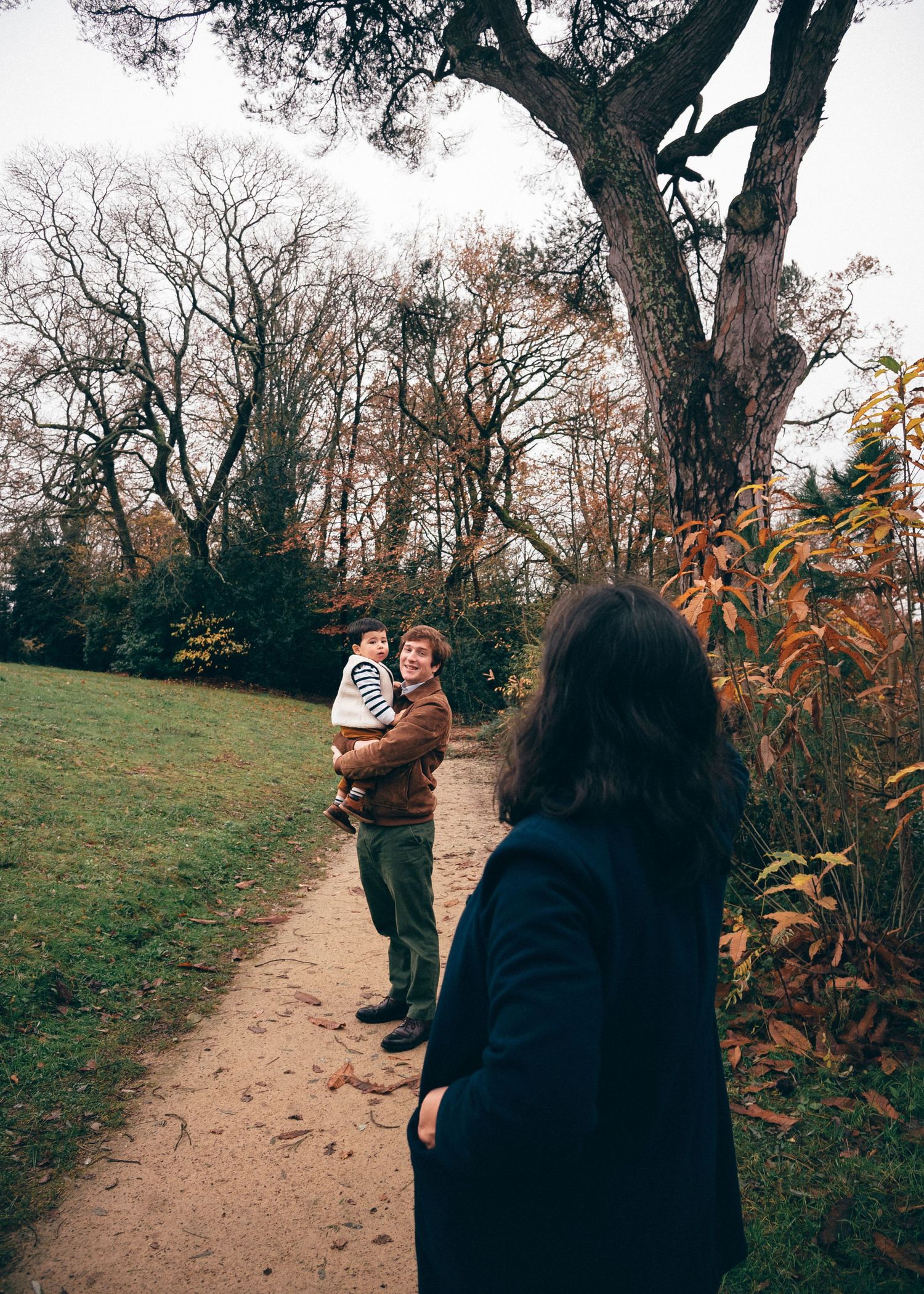 Scène de famille dans un parc arboré, regard porté sur un parent tenant son enfant lors d’une séance photo à Nantes