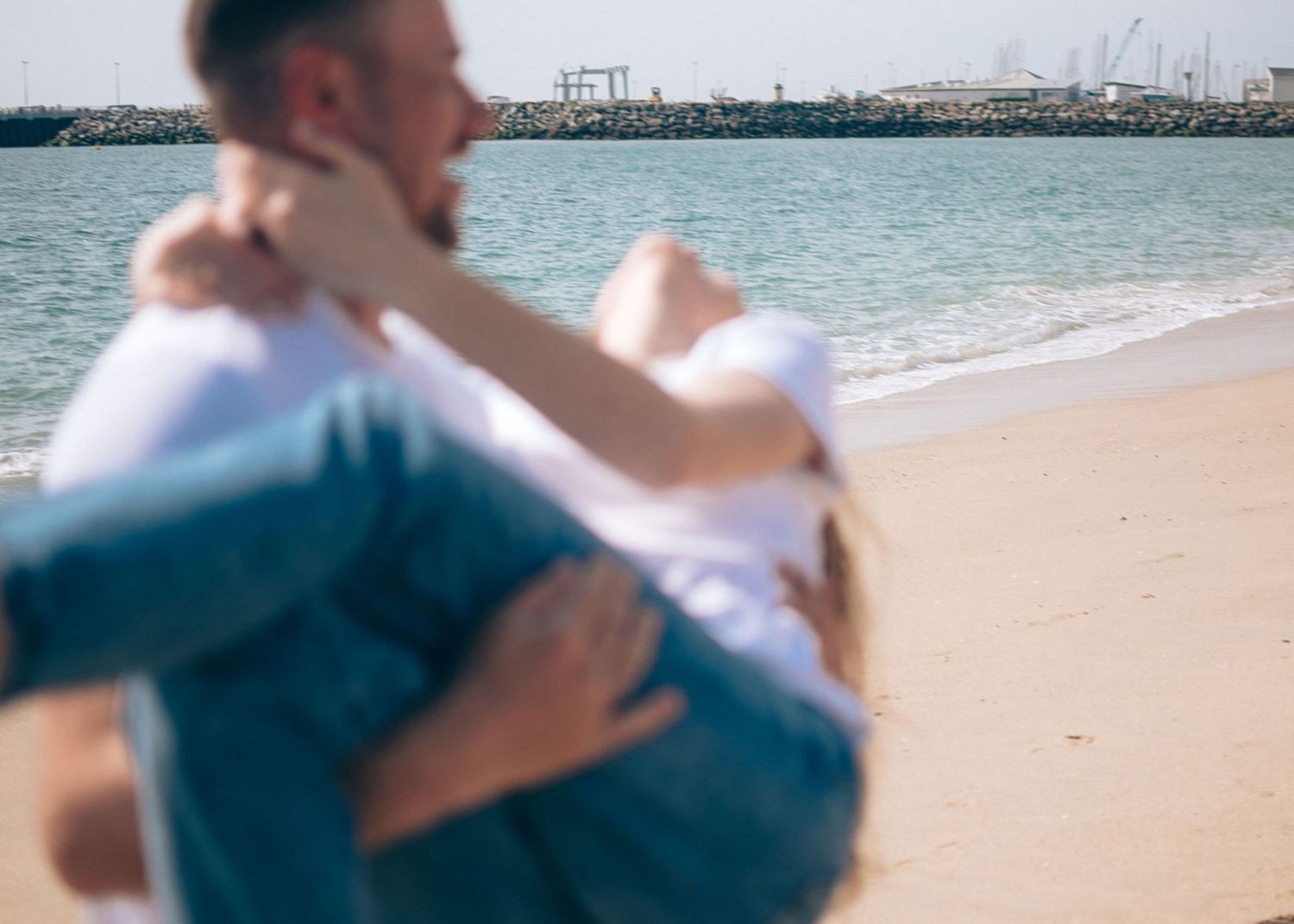 Two people embrace playfully on a sandy beach with a harbor visible in the distance.
