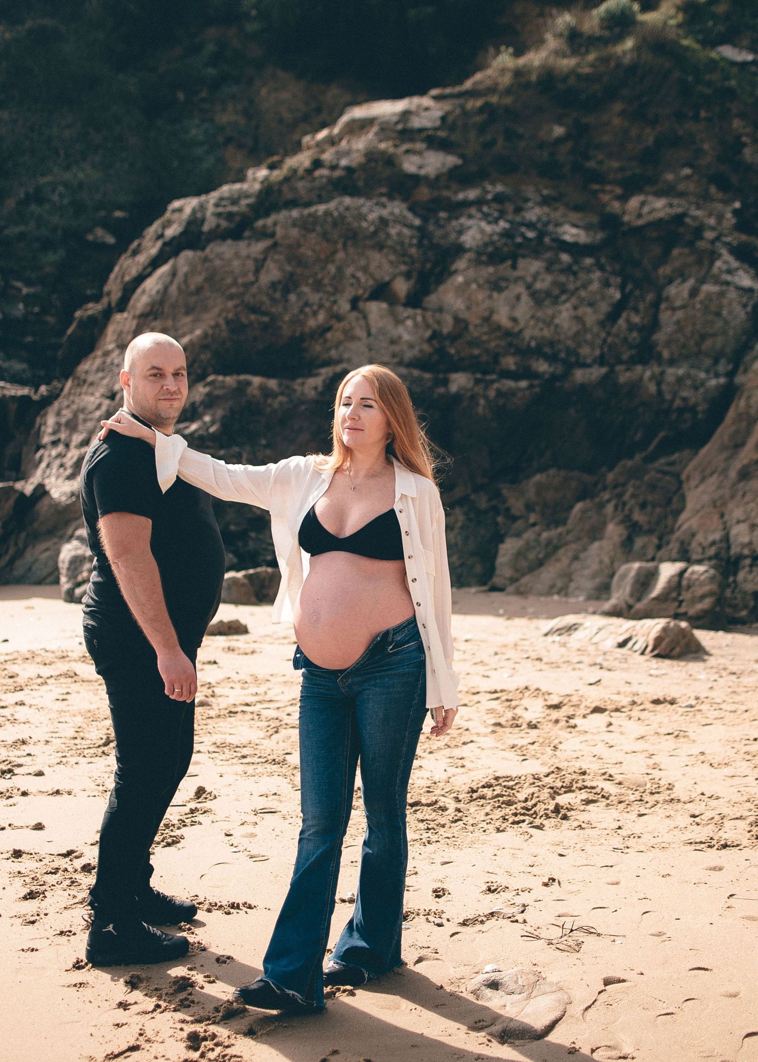 A couple shares a tender moment during a beachside maternity photoshoot near rocky cliffs.