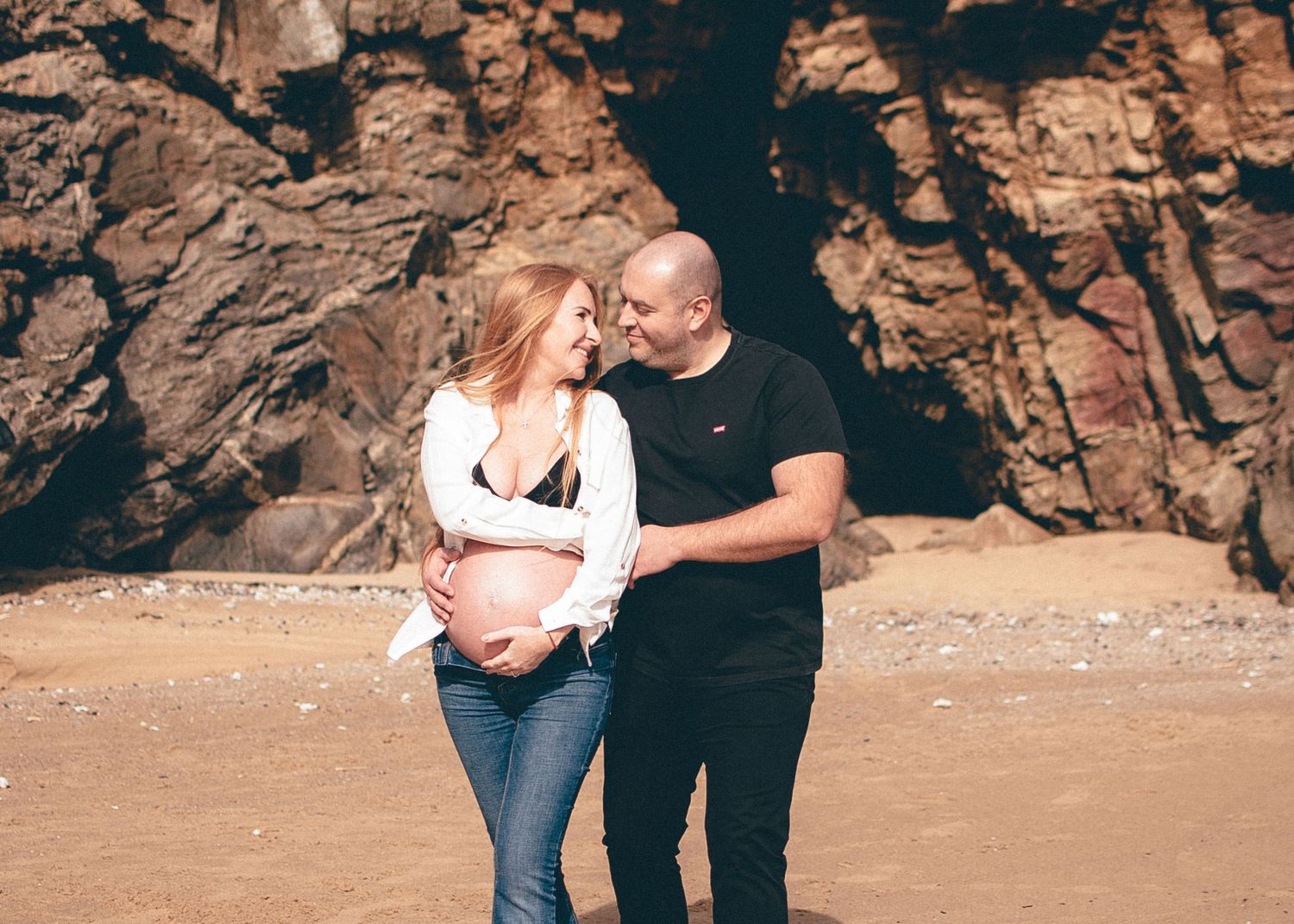 A maternity photography session unfolds on a sandy beach with dramatic rock formations.