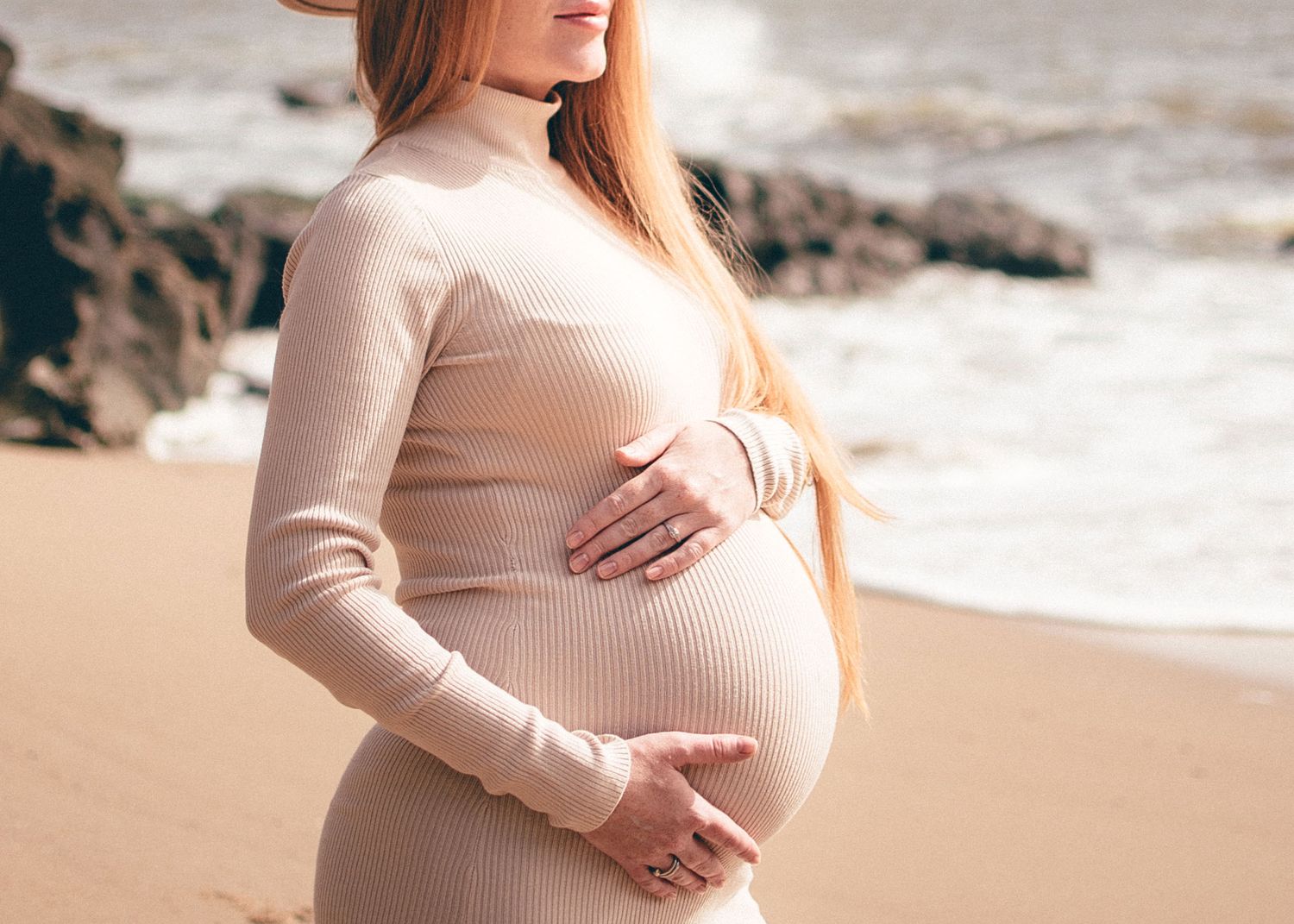 A person in a beige sweater poses on a beach during a maternity photoshoot.