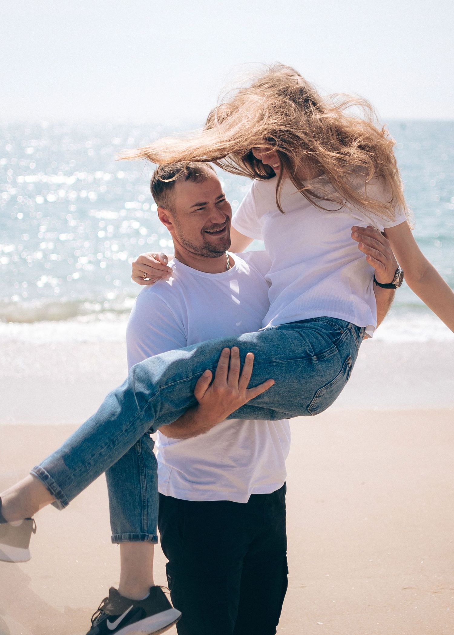 Two people in casual attire share a playful dance moment on a sunny beach.
