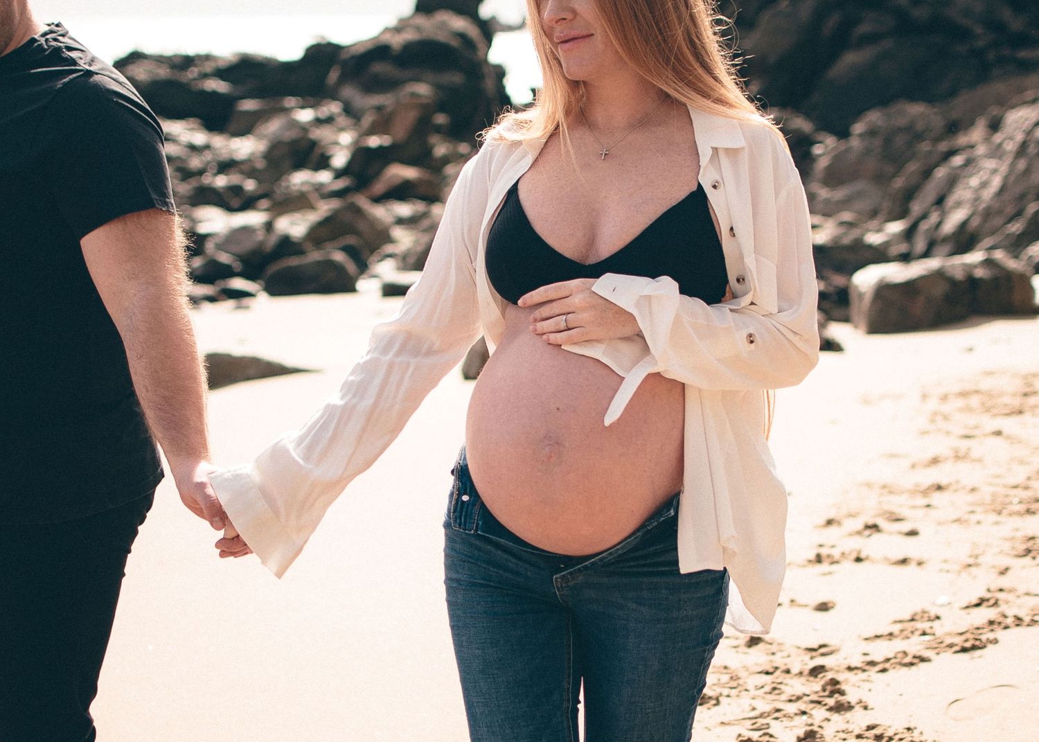 A maternity photo session on a beach with dramatic rocky cliffs in the background.