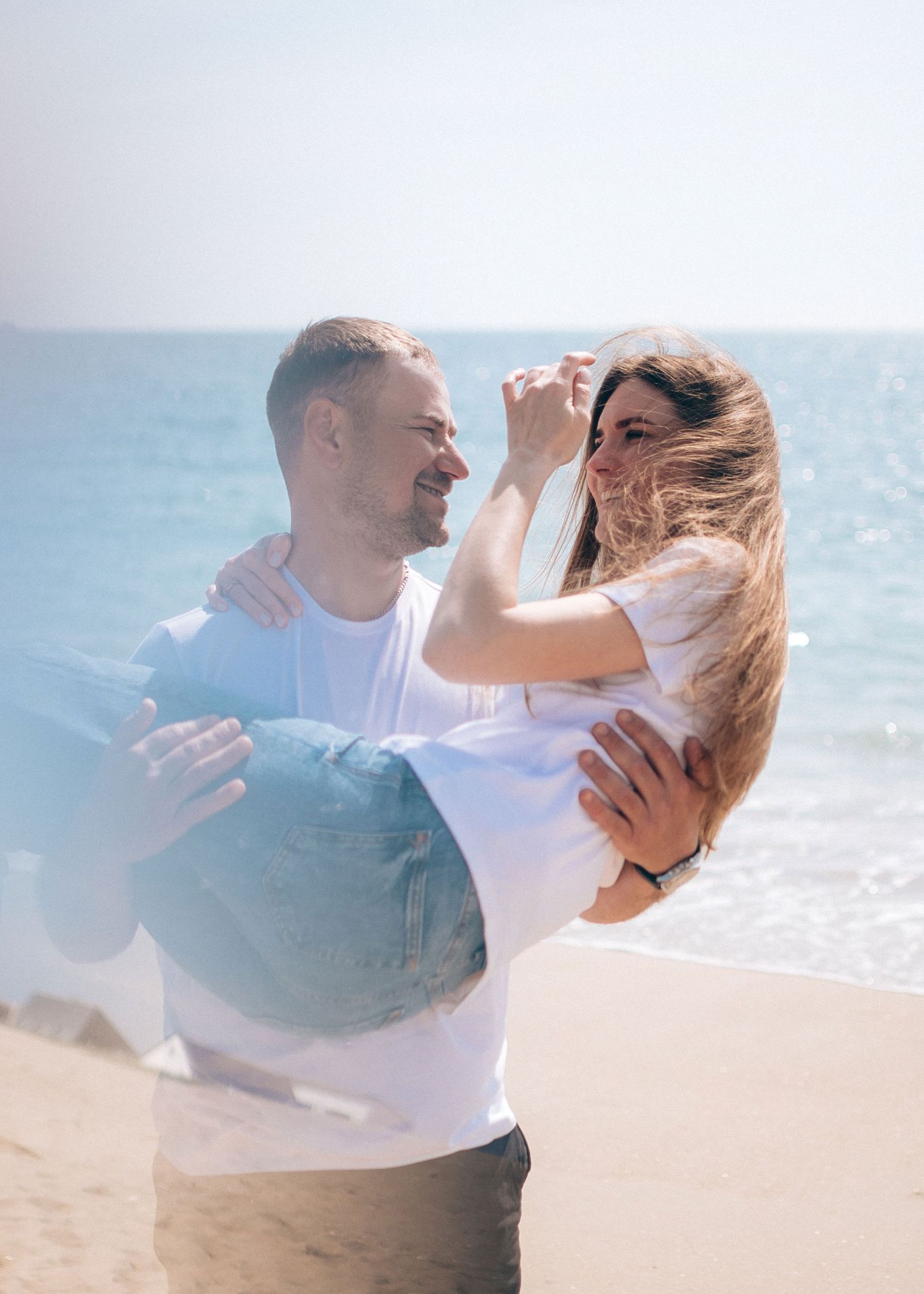 A couple in white and blue outfits dances joyfully on a beach with waves crashing behind them.