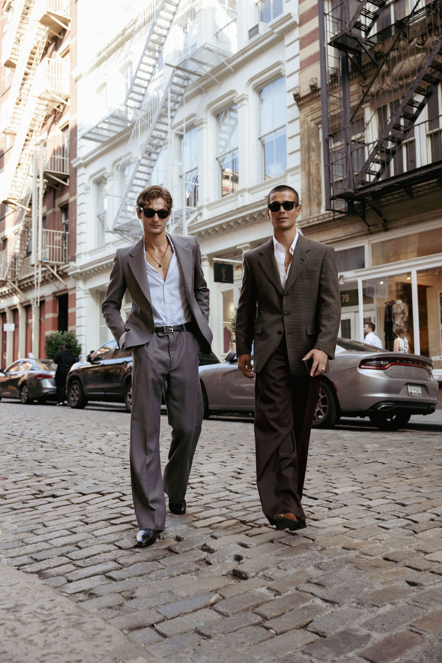 Two men in suits walking down a cobblestone street in an urban setting with historic buildings in the background.