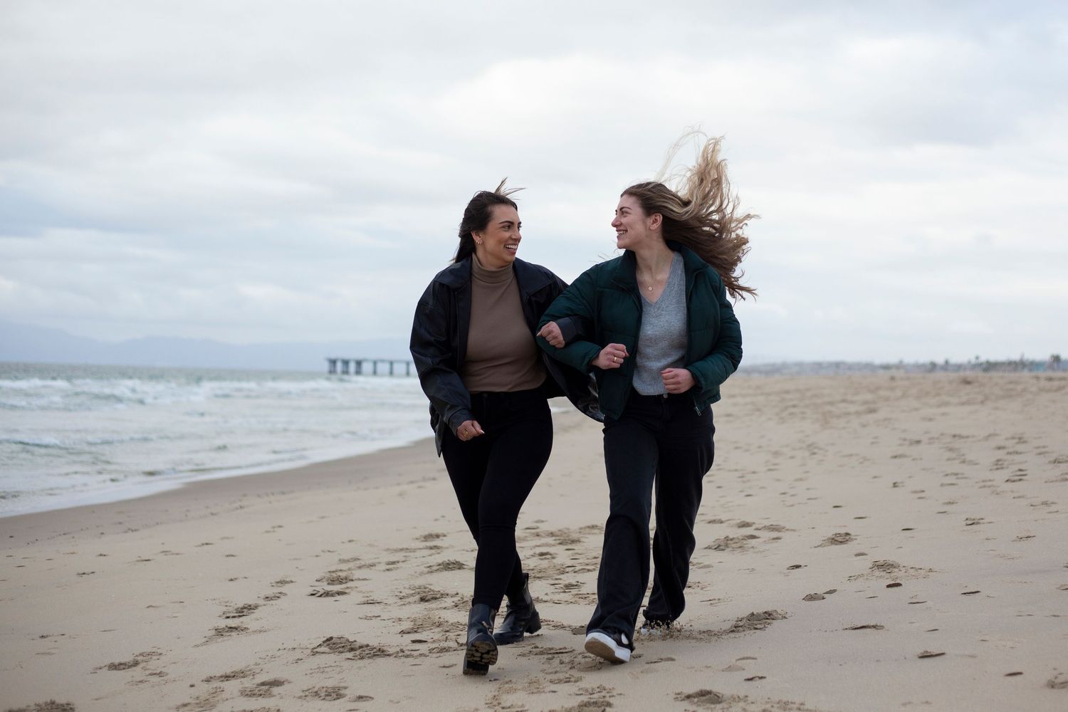 Two sisters walking together on a sandy beach during an overcast day with waves in the background.
