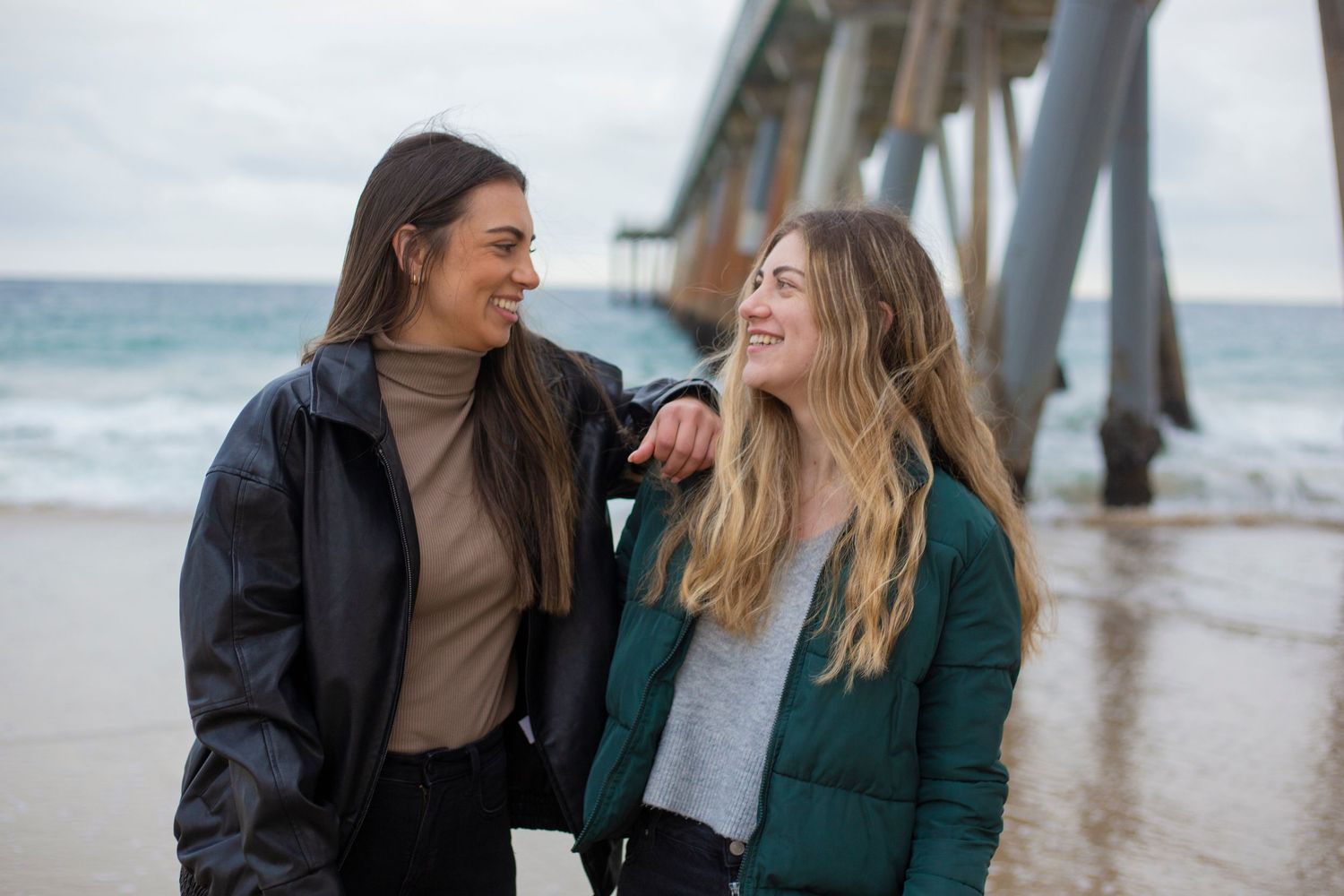 Two friends share joyful laughter while sitting together on a beach pier during an overcast day.