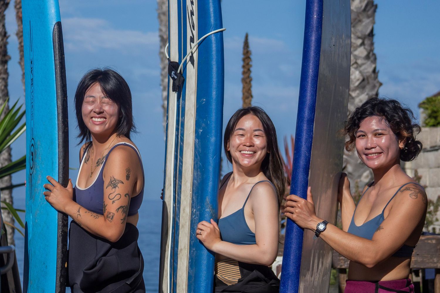 A sequence of joyful friends posing and laughing between bright blue surfboards outdoors on a sunny day.